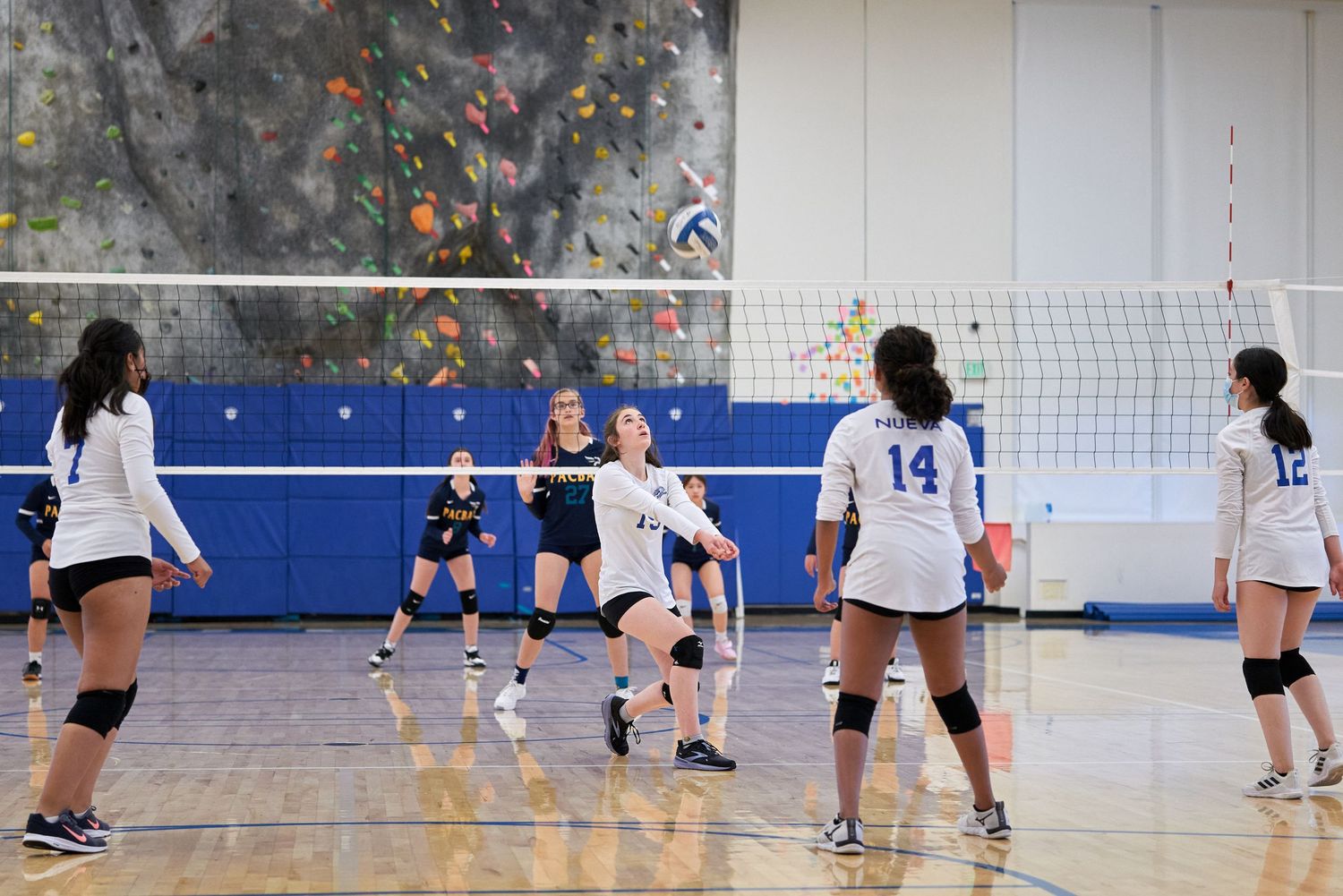 Indoor volleyball match being played on court with large climbing wall visible in background.