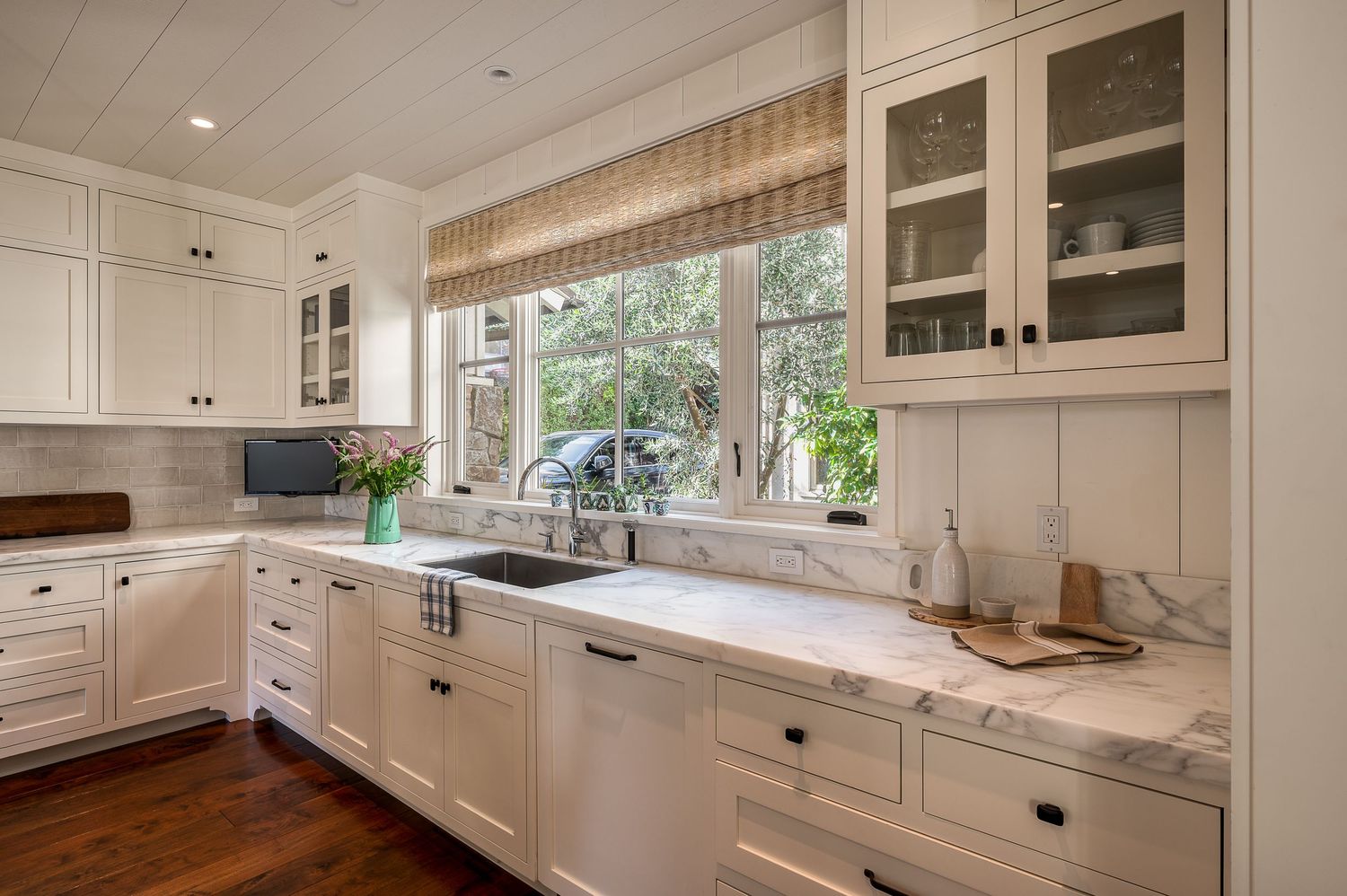 Modern white kitchen with marble countertops, glass-front cabinets, and large window overlooking outdoor greenery.