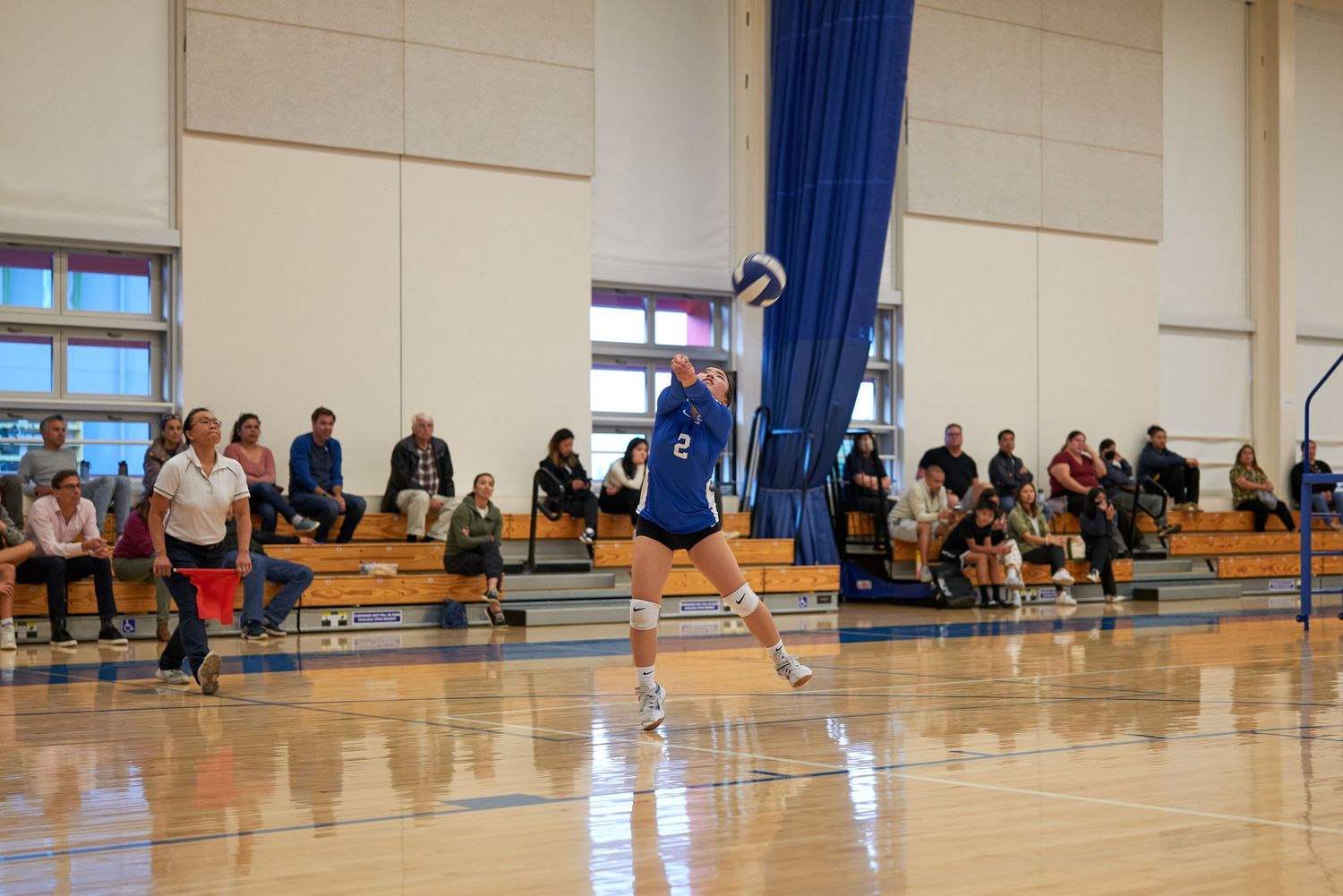 A volleyball player serves during a match while spectators watch from the bleachers.