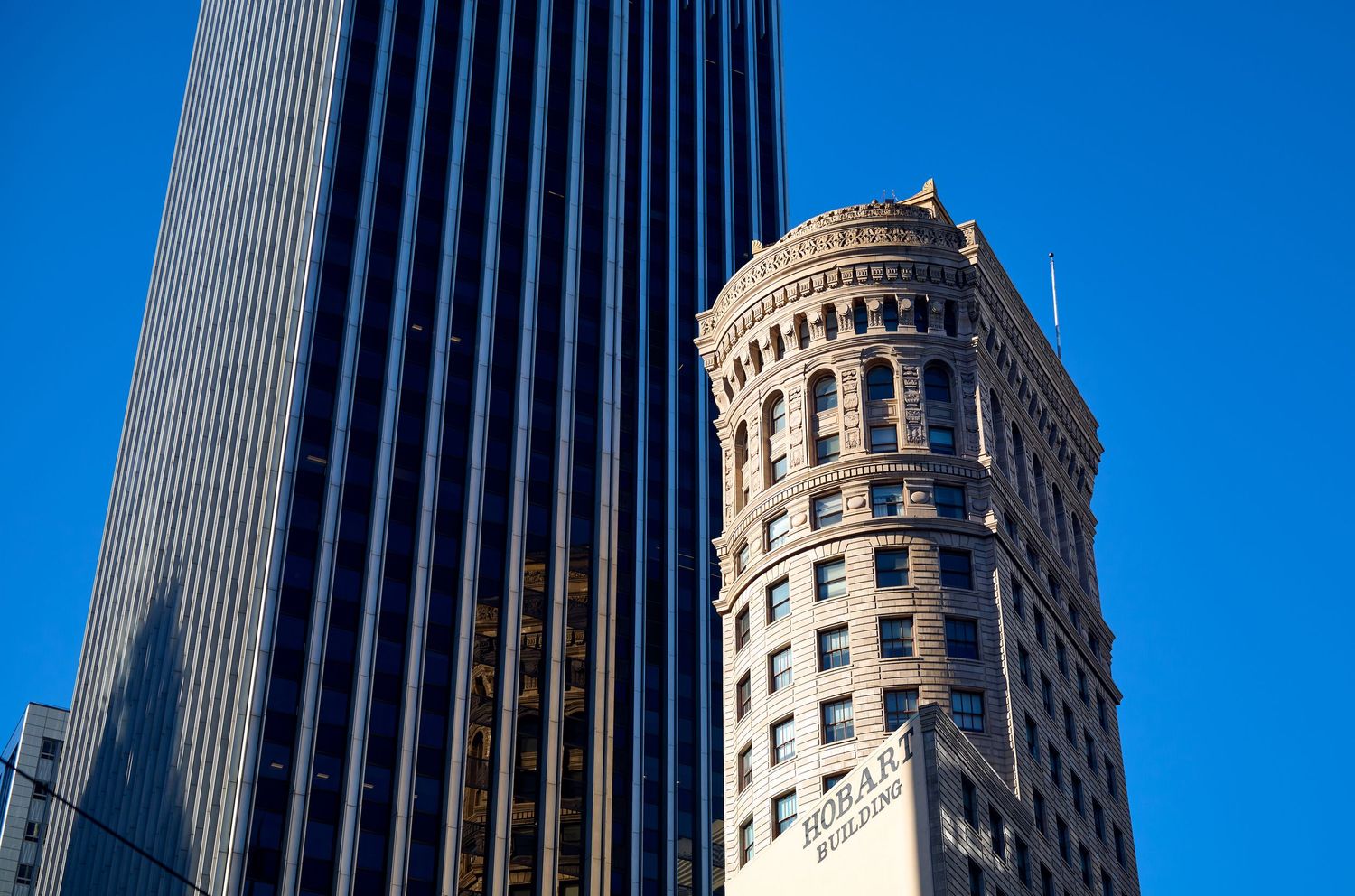 Modern glass skyscraper contrasts with historic circular tower building against bright blue sky.