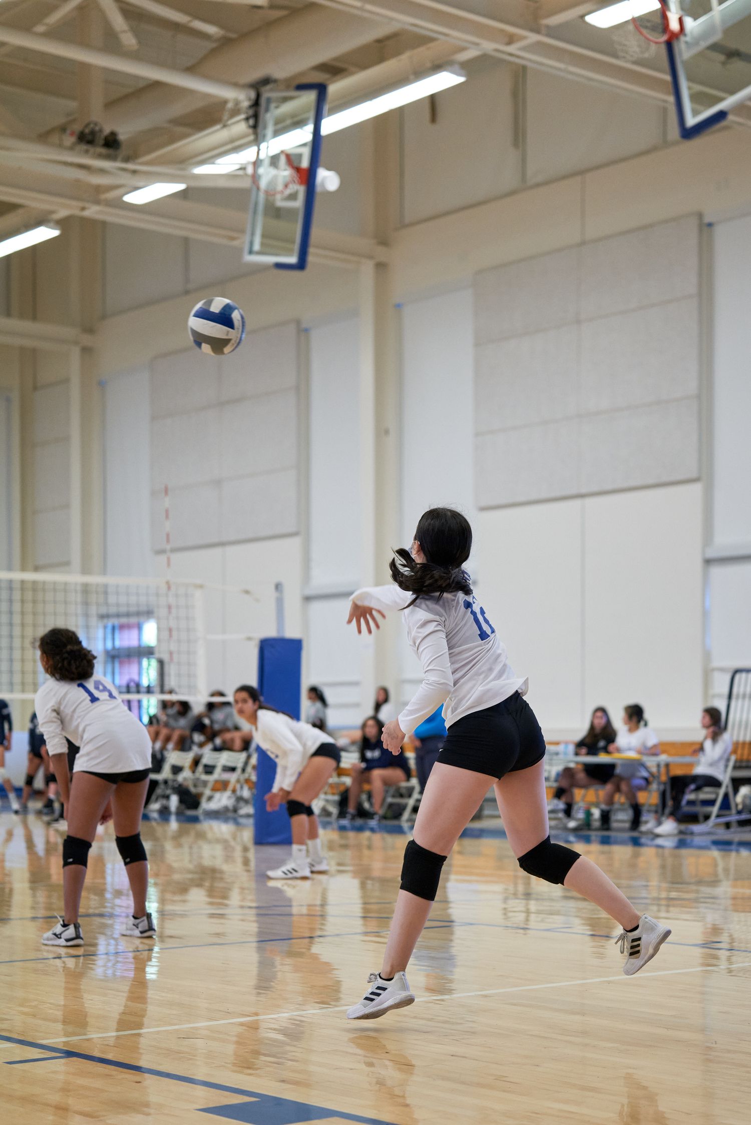 Player leaping to serve volleyball during indoor match with net and court visible.