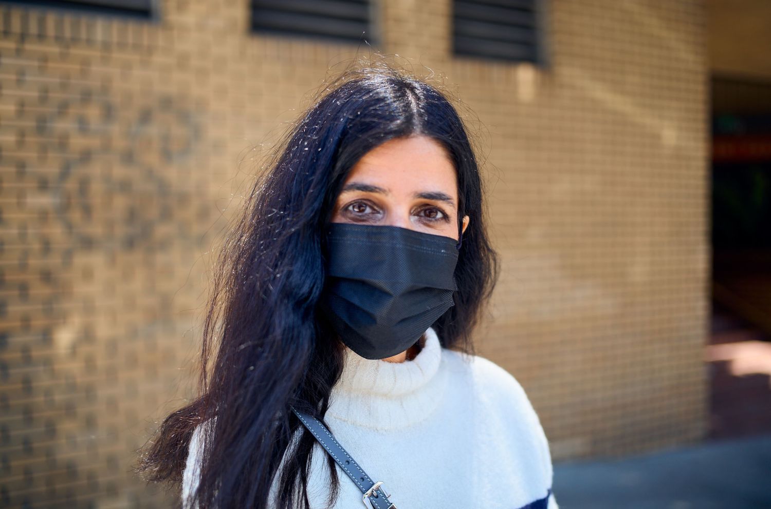 Person with long dark hair wearing white shirt and black face mask stands against brick wall.