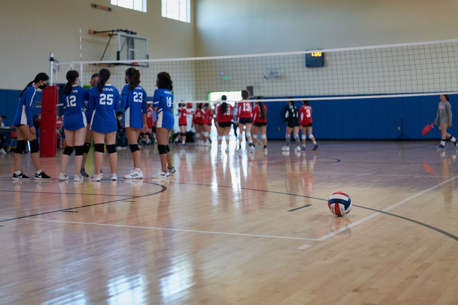 Volleyball players from opposing teams line up on either side of the net during a match in an indoor gymnasium.
