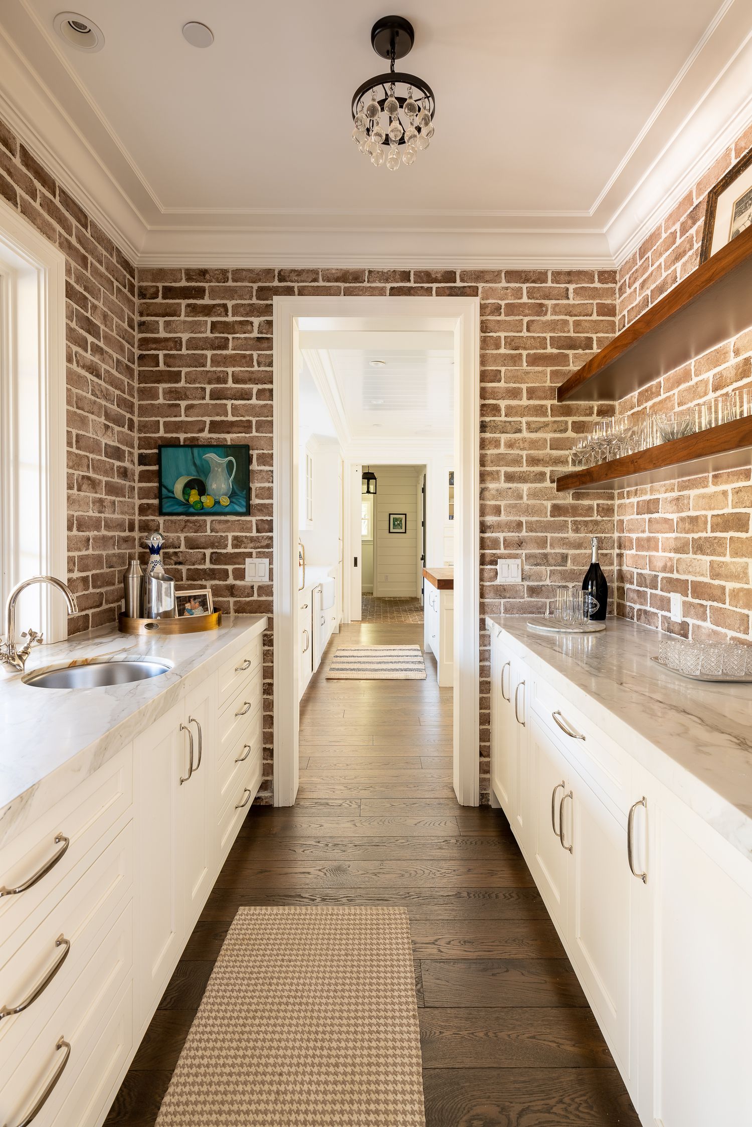 Modern galley kitchen with exposed brick walls, white cabinets, and wooden floating shelves features a crystal chandelier.