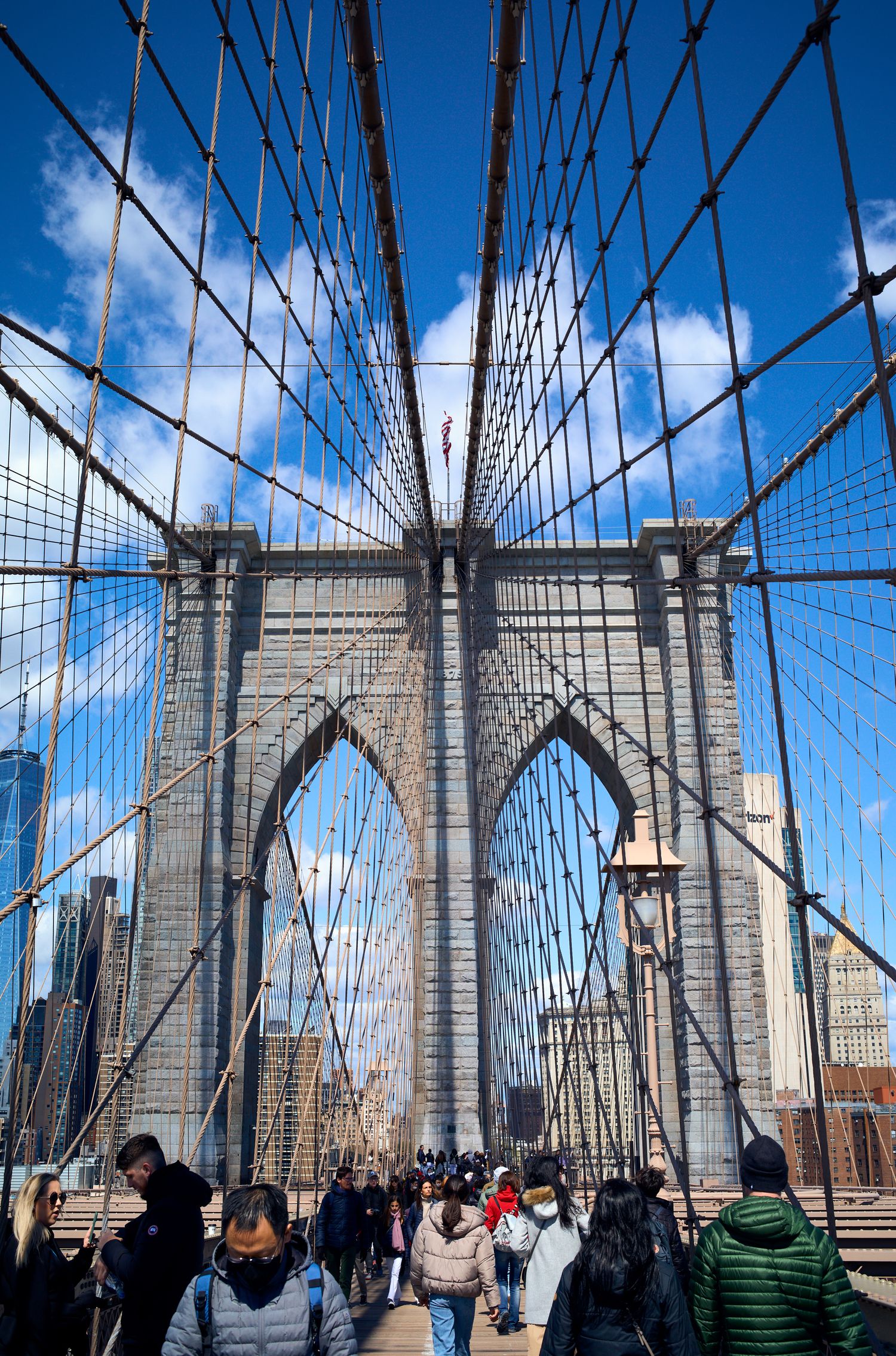 View of architectural cables and stone towers of Brooklyn Bridge with crowds walking across on a sunny day.