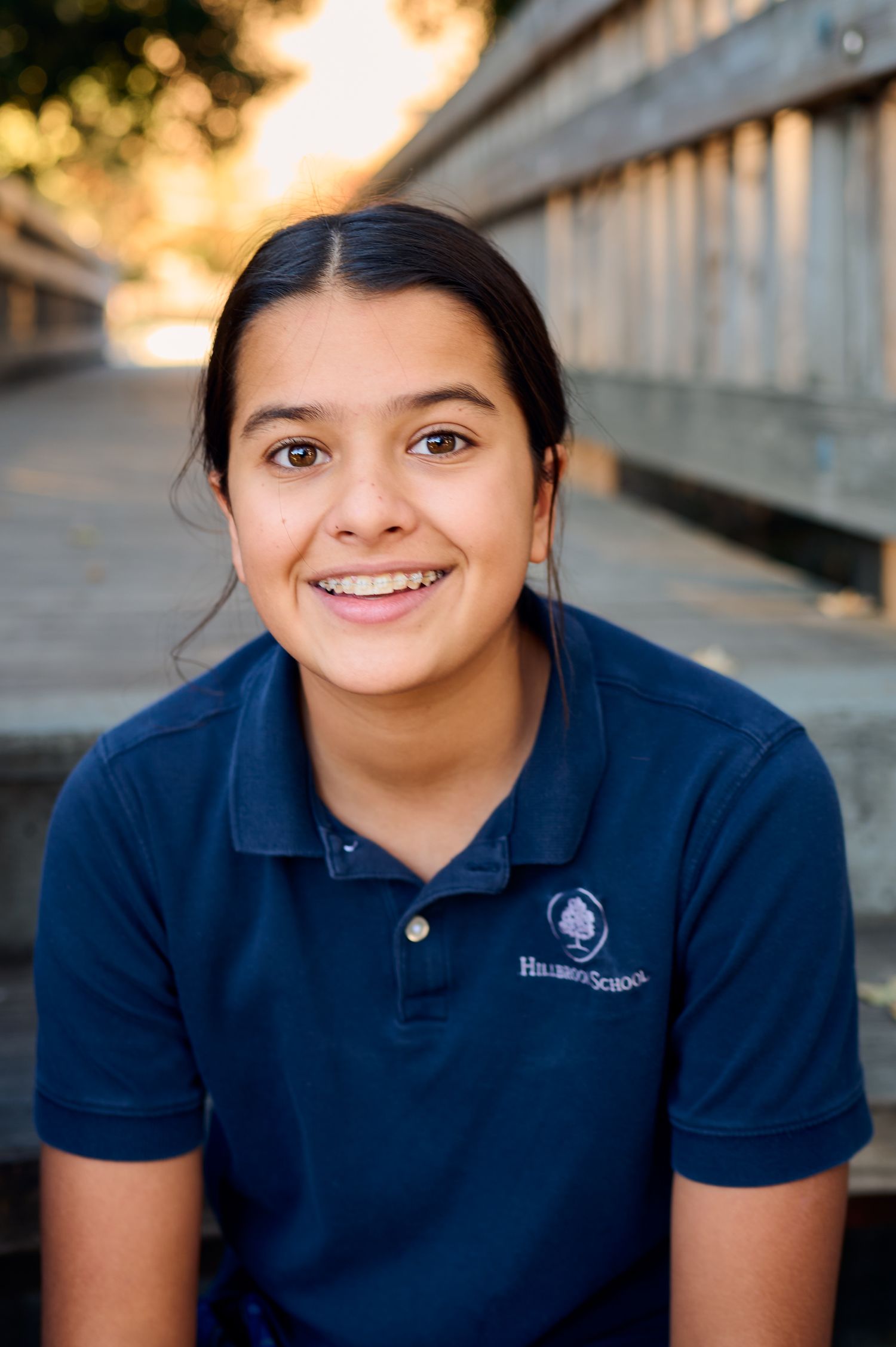 Student in a dark blue polo shirt with an embroidered logo smiles outdoors during golden hour.