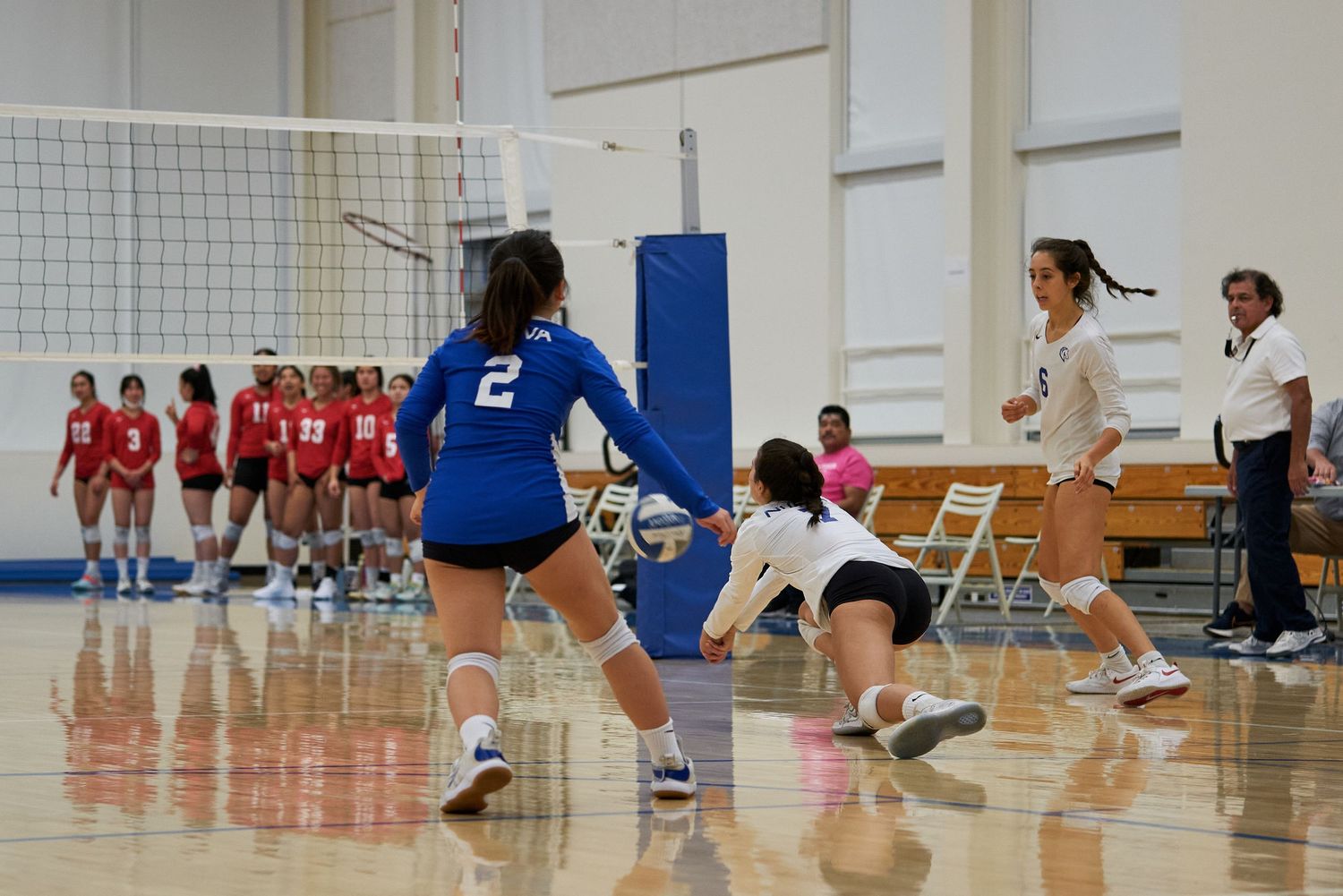 Players in blue and red uniforms compete during a volleyball match in a gymnasium.