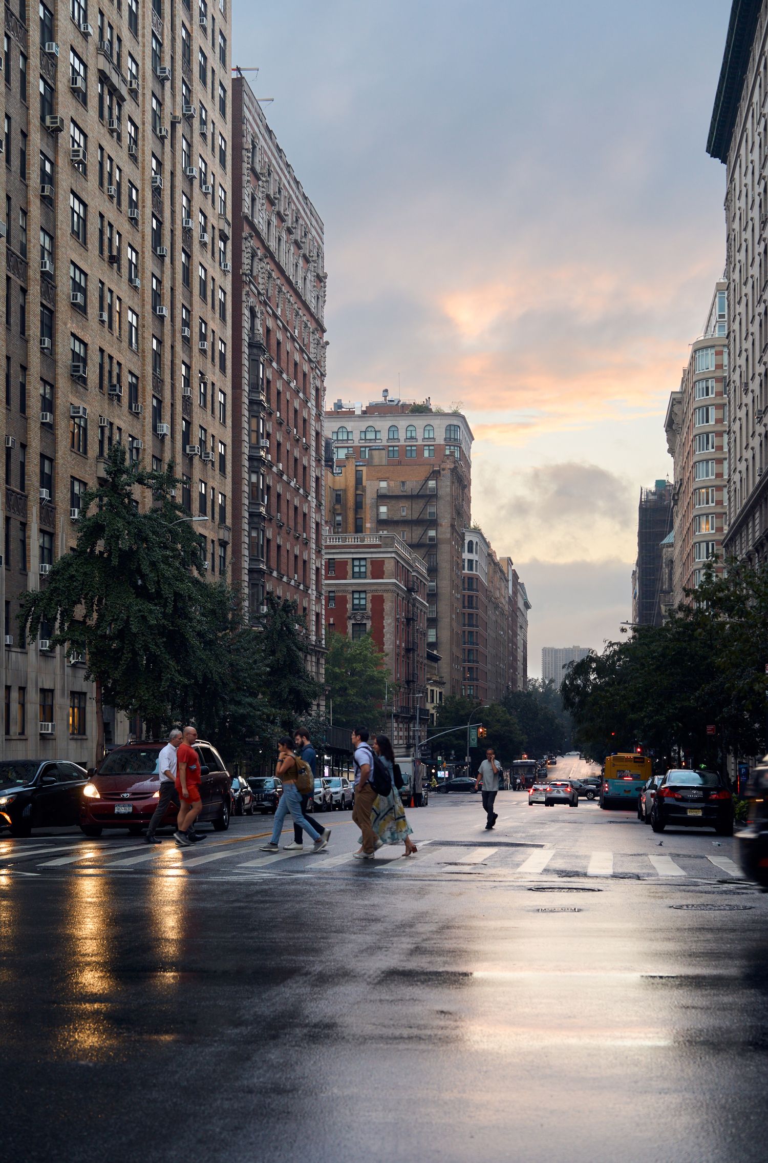 Rainy city street at sunset with tall apartment buildings and reflective wet pavement.