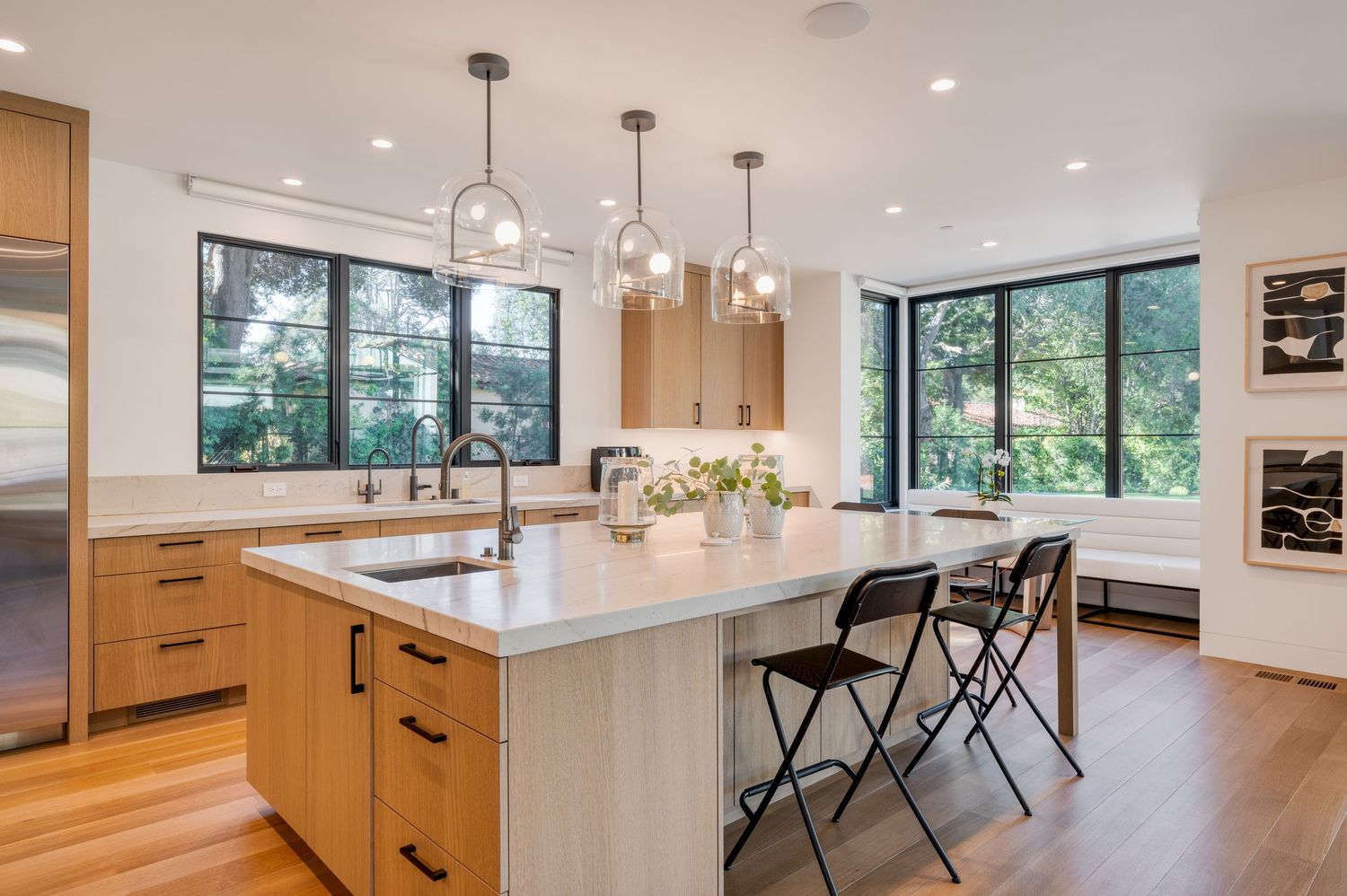 Contemporary kitchen with wood cabinets, white island, and floor-to-ceiling windows overlooking greenery.