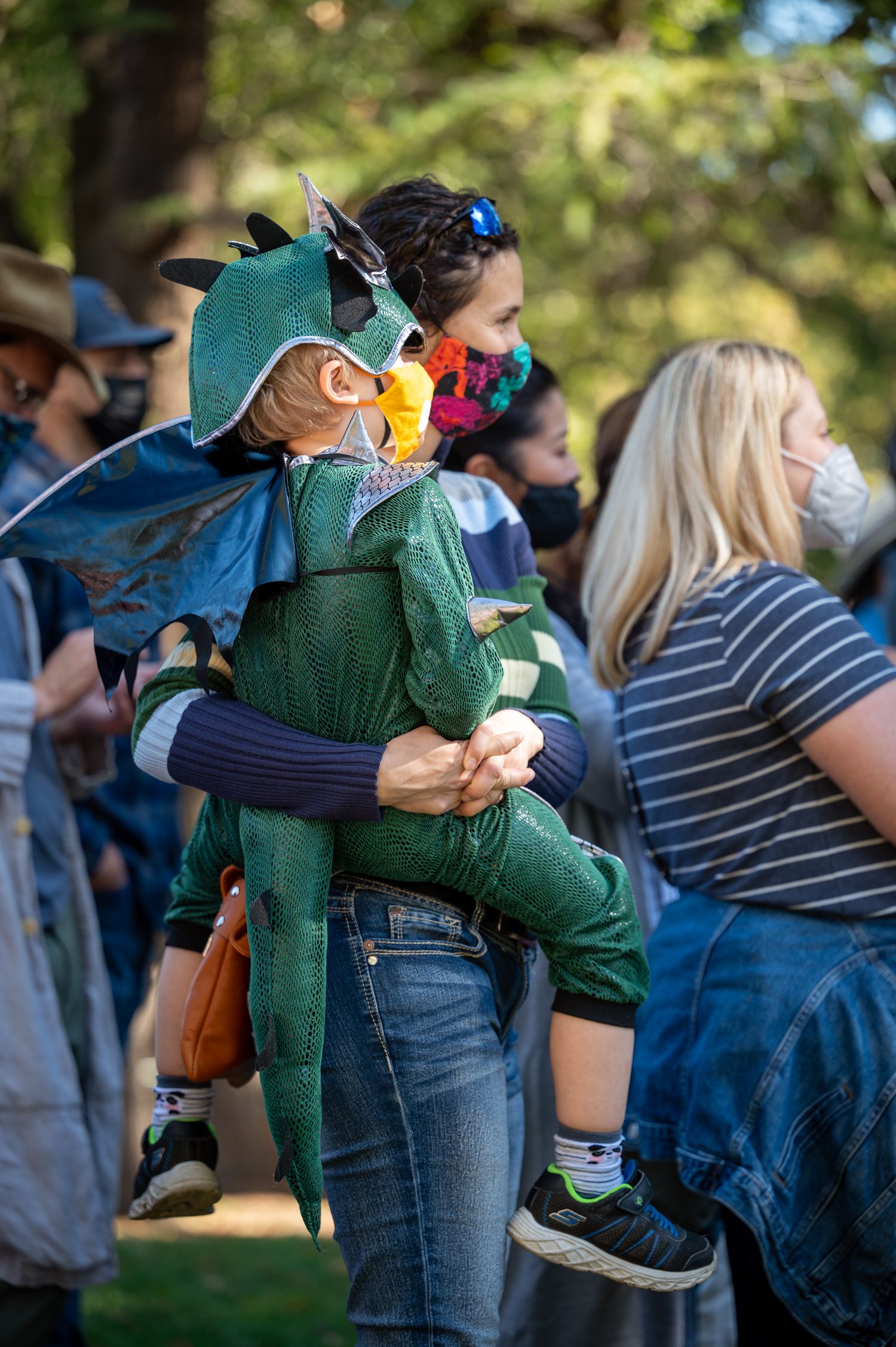 Person carrying a child in dinosaur costume at an outdoor gathering.