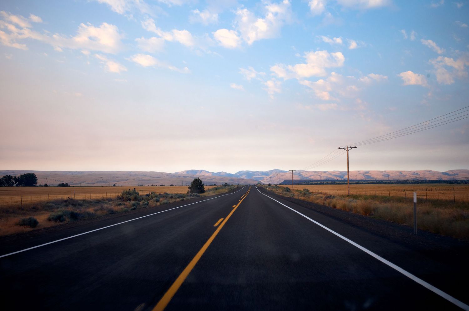 Empty highway stretches towards the horizon through rural countryside under partly cloudy skies.