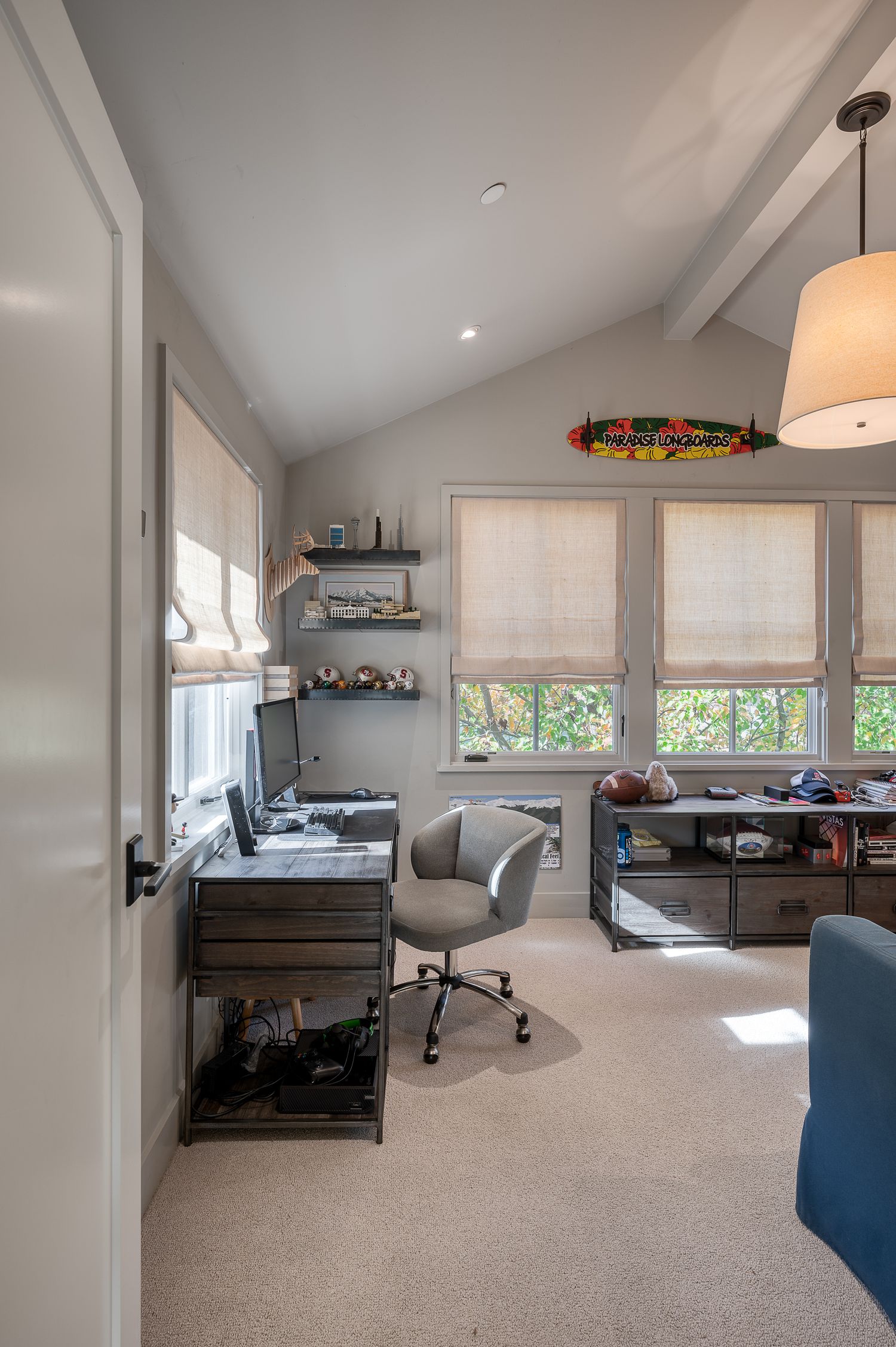Modern home office with sloped ceiling, window shades, floating shelves and ergonomic desk setup.