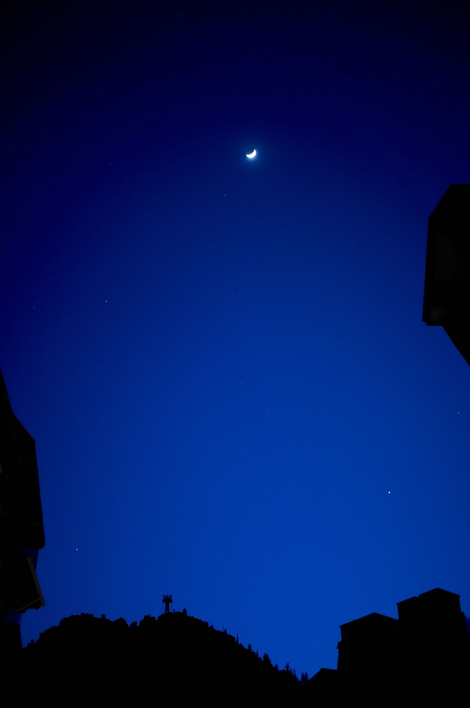 Crescent moon shines in deep blue night sky above dark silhouetted buildings and landscape.