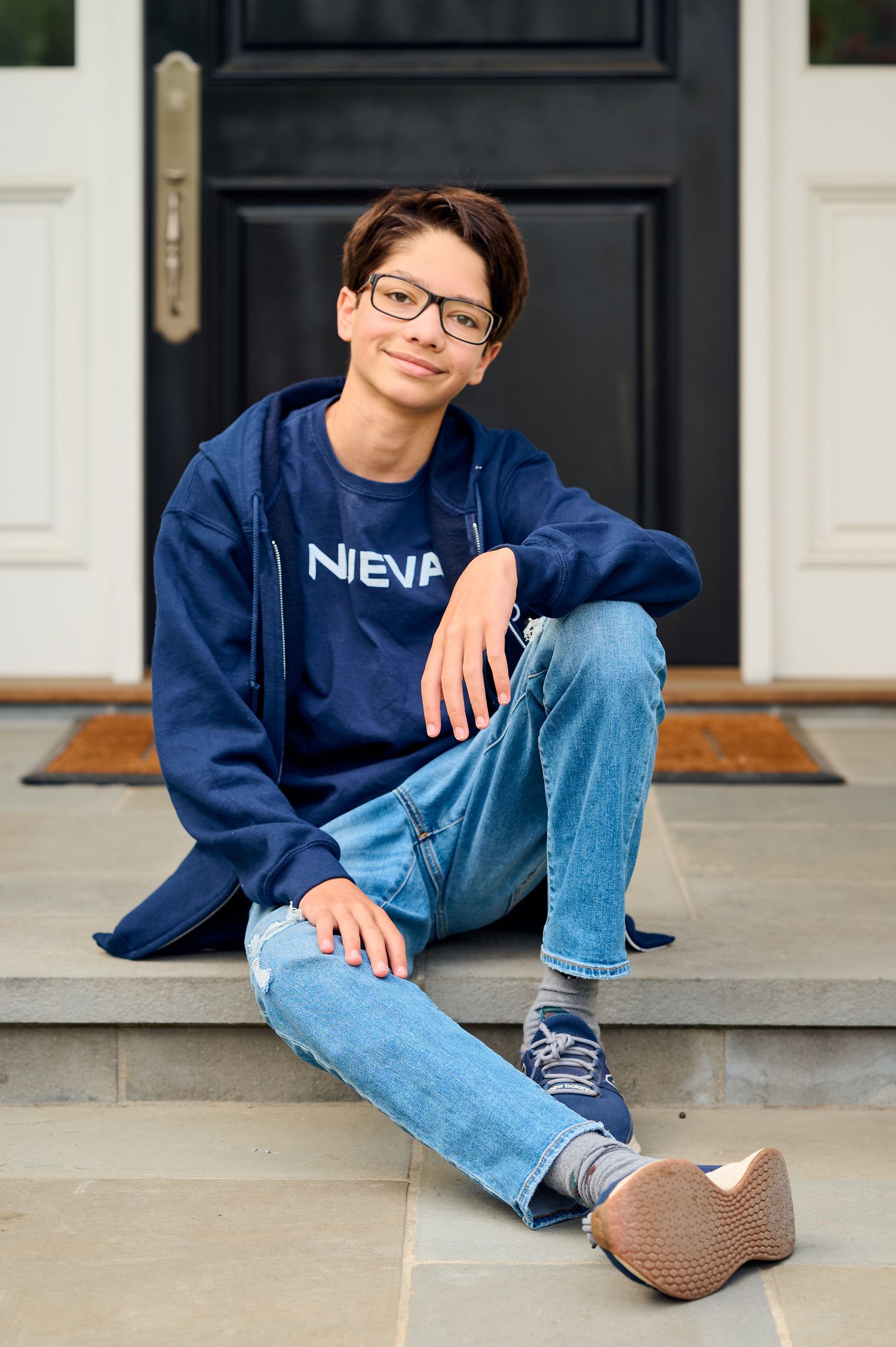Person in navy NEVA sweatshirt and jeans sits casually on front porch steps.
