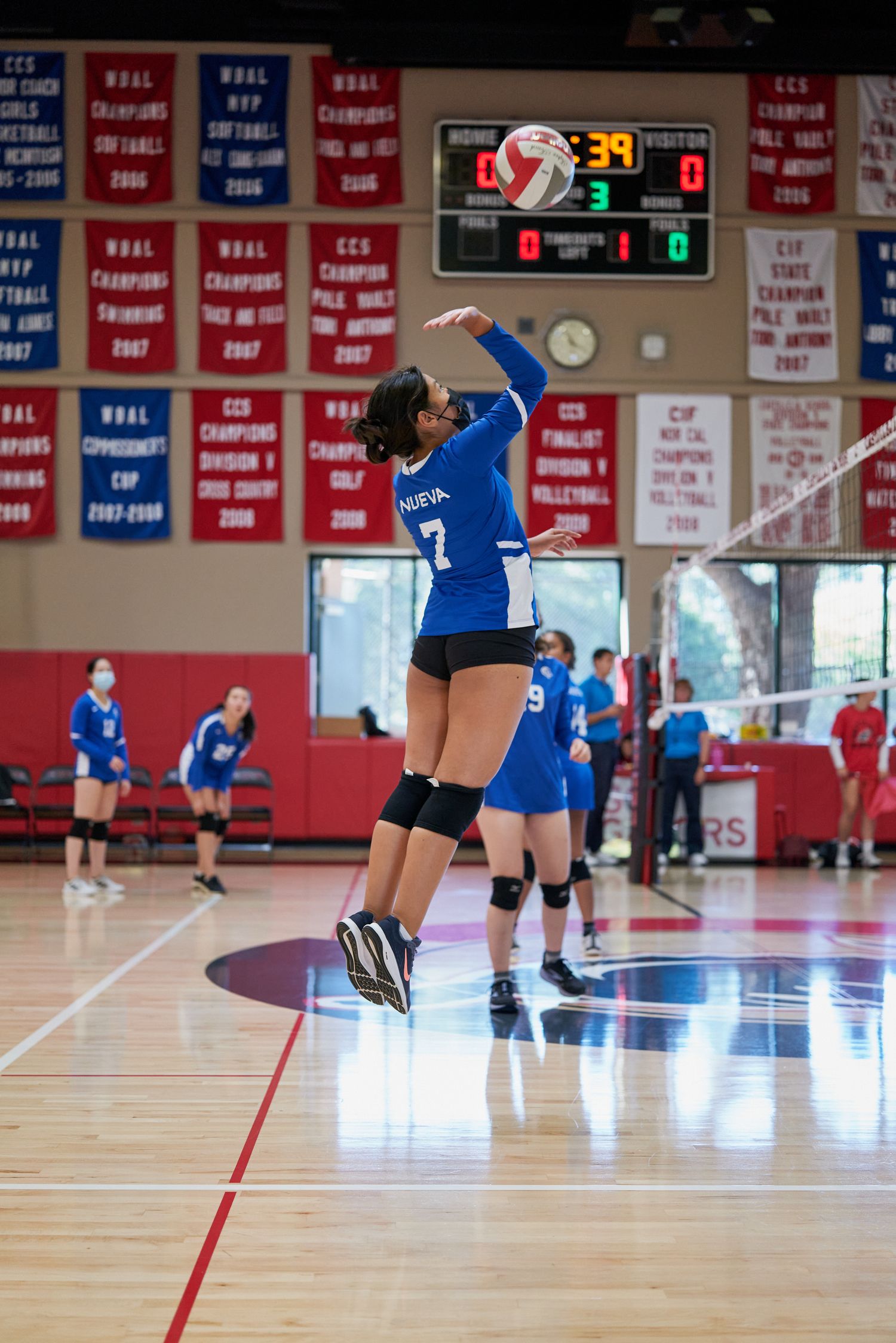 Player in blue jersey jumps to serve volleyball during competitive match in gym with championship banners.