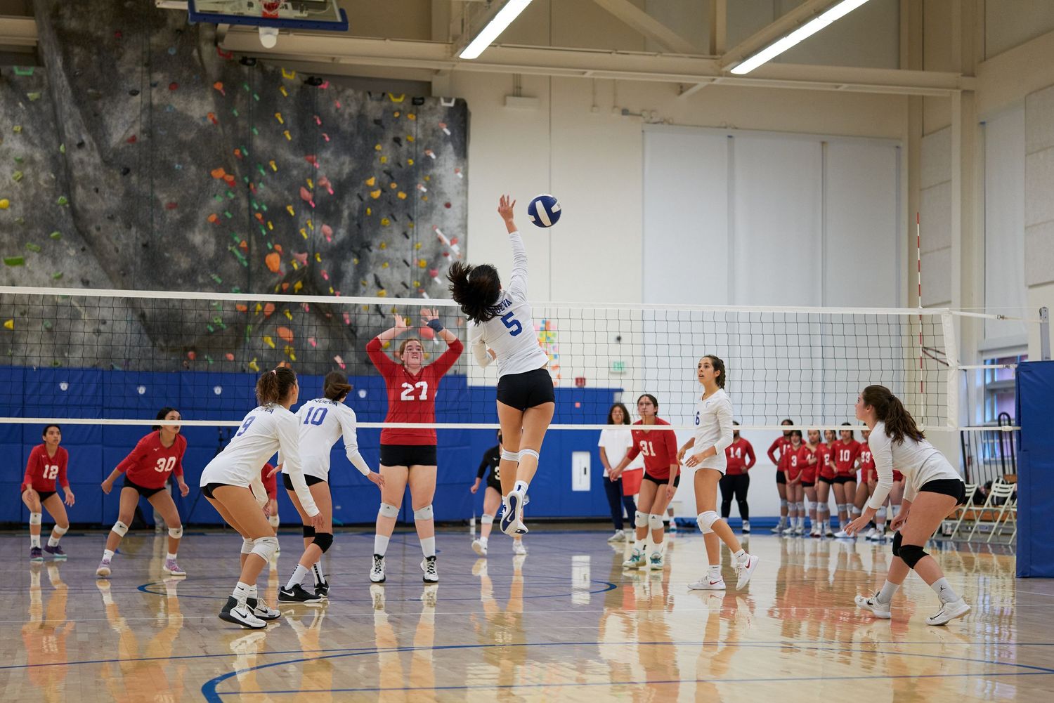 Two teams compete in an indoor volleyball match with players jumping at the net.
