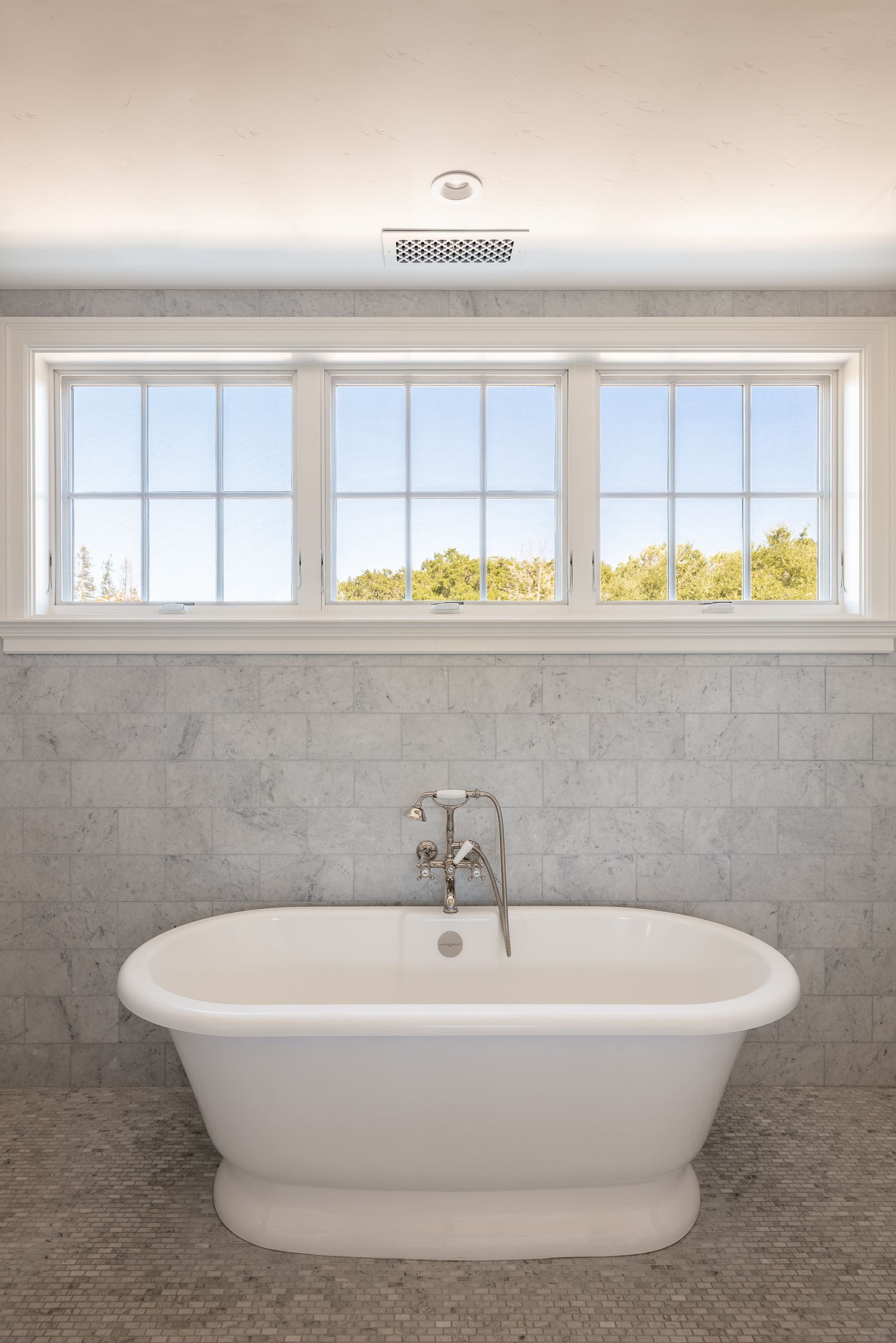 Freestanding white bathtub beneath three windows offering natural light in a luxury bathroom.