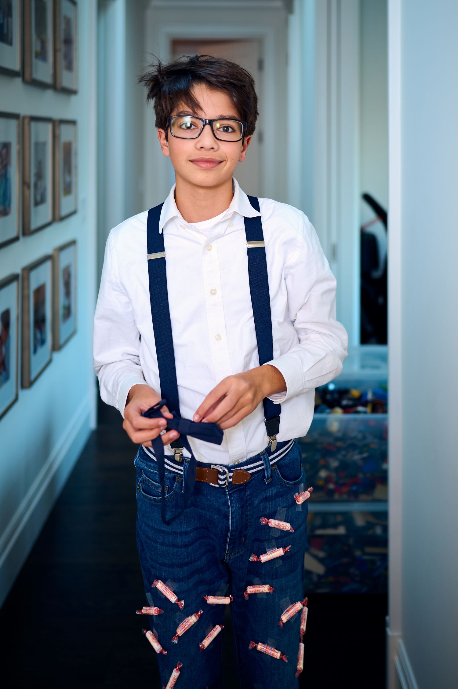 A person wearing suspenders and a white button-down shirt stands in a hallway with framed photos.
