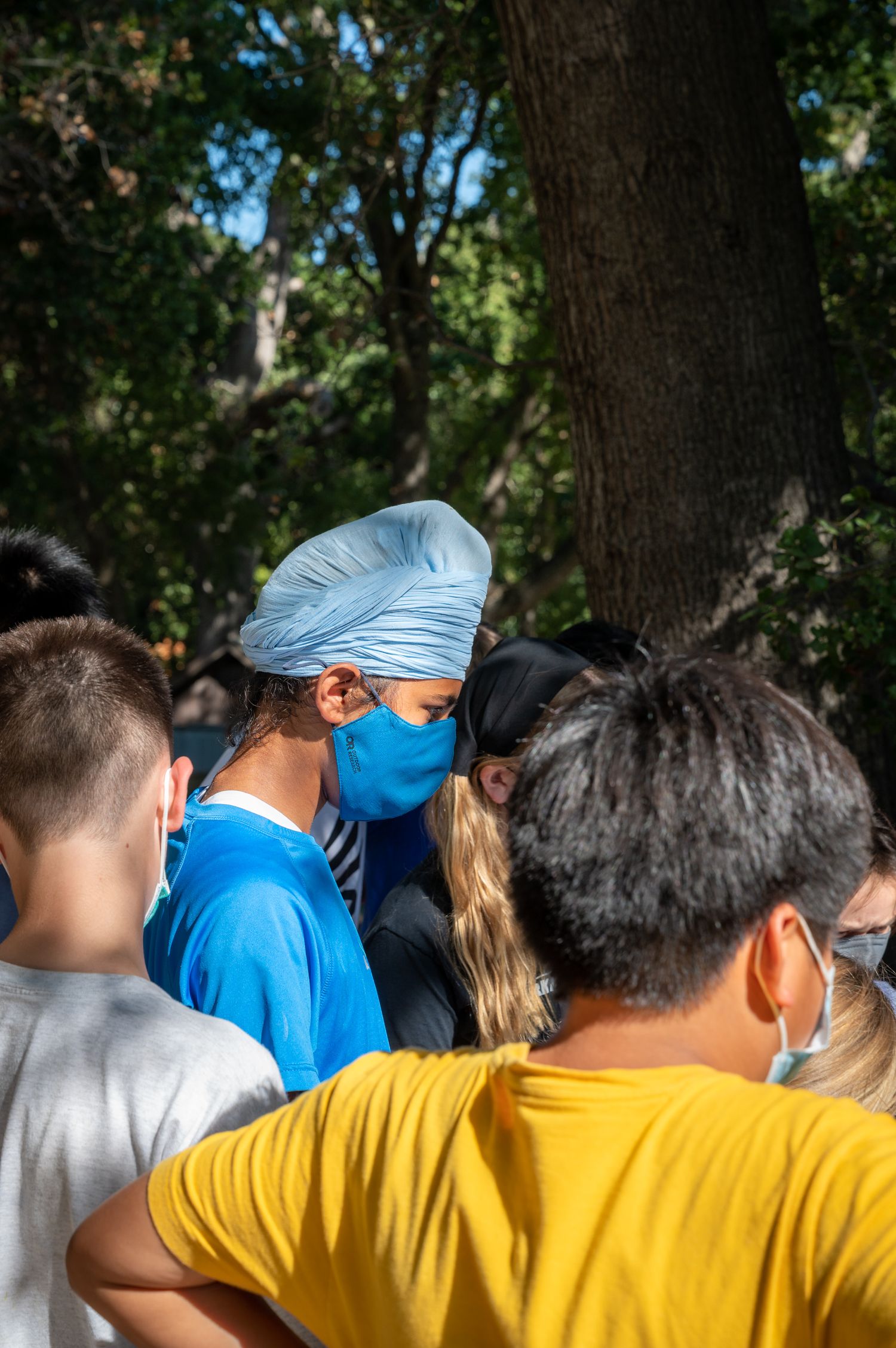 People in face masks stand together outdoors under tree cover.