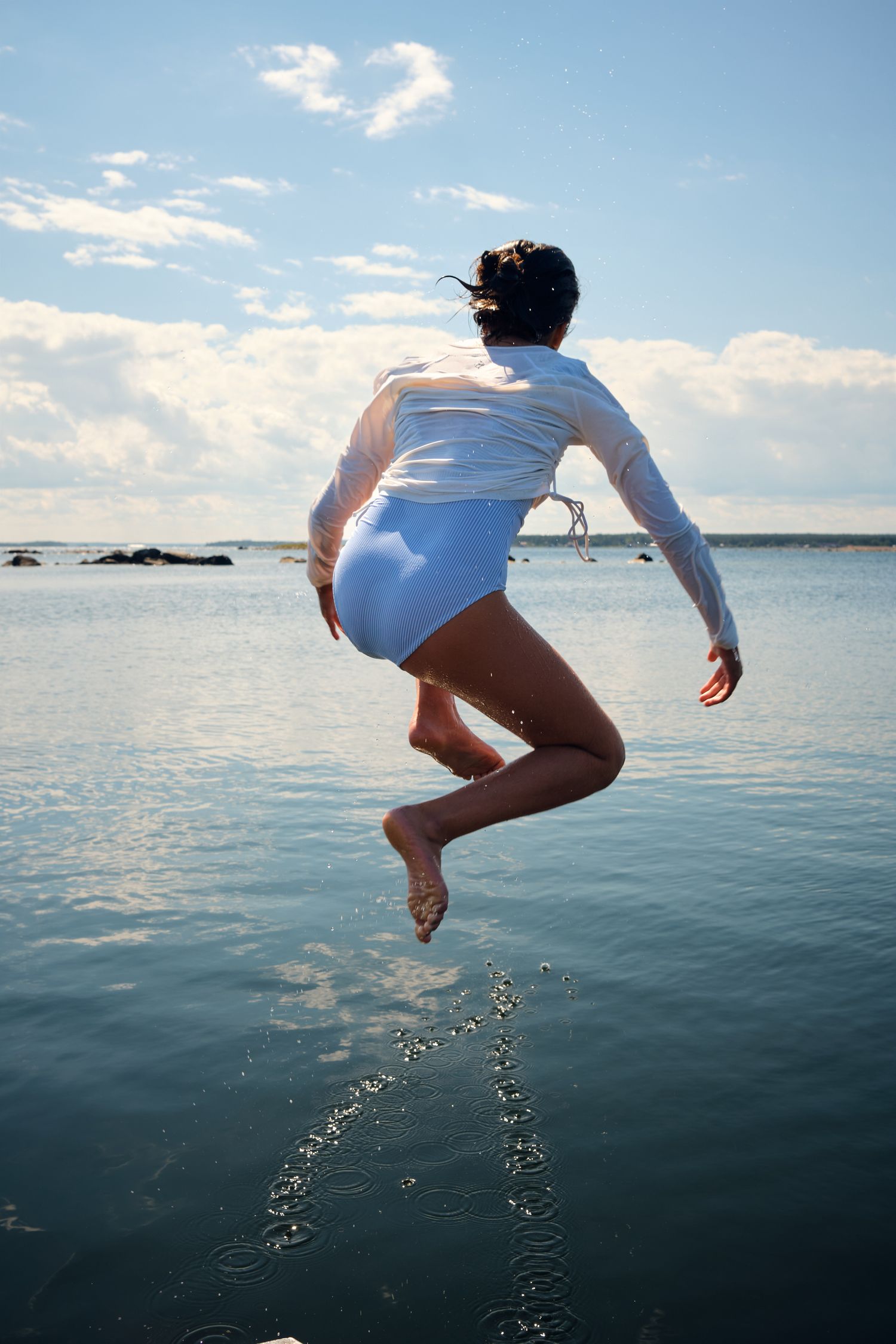 Person in white swimsuit jumping into water against blue sky with clouds.