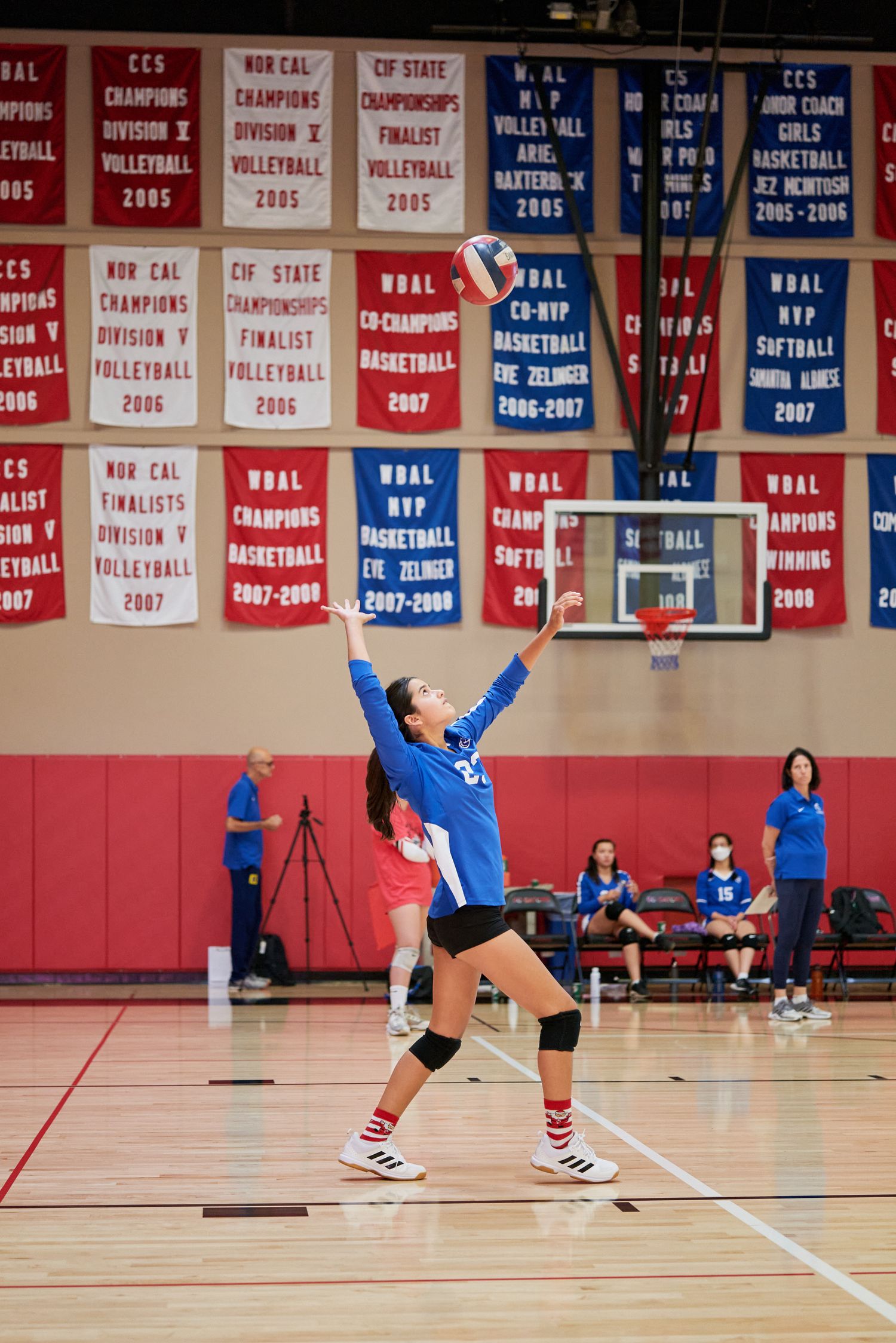 Volleyball player serving ball against wall of championship banners in school gymnasium.
