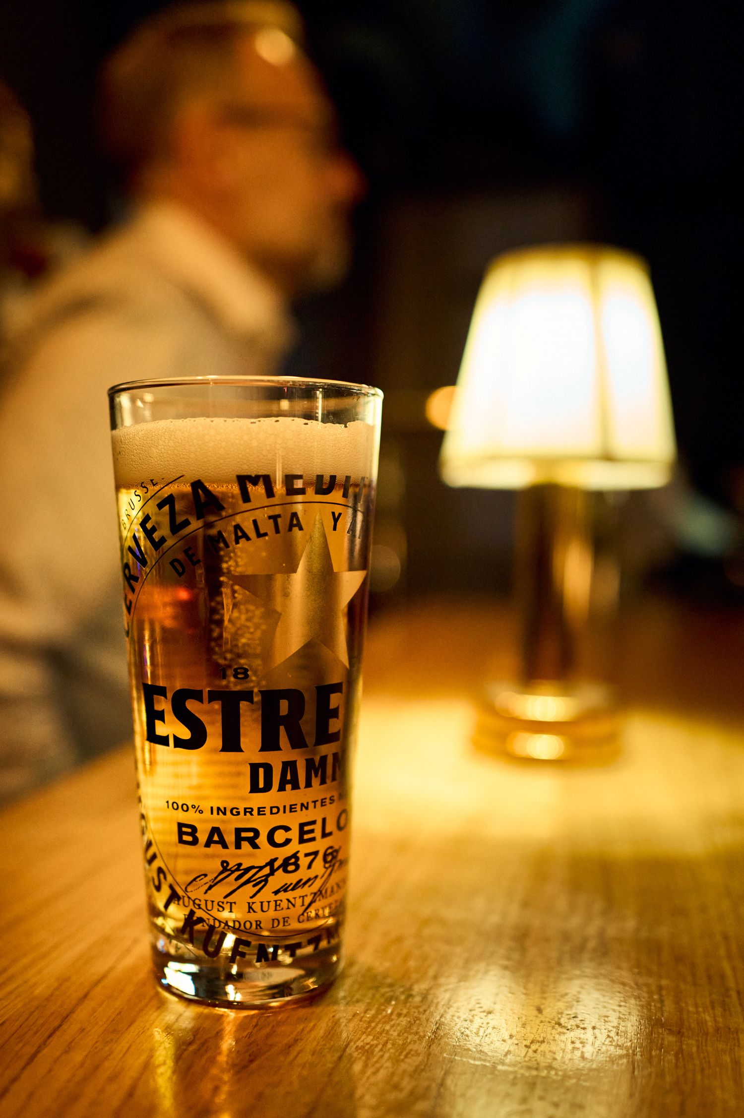 A glass of Estrella Damm beer from Barcelona on a wooden table with a warm lamp glow in the background.