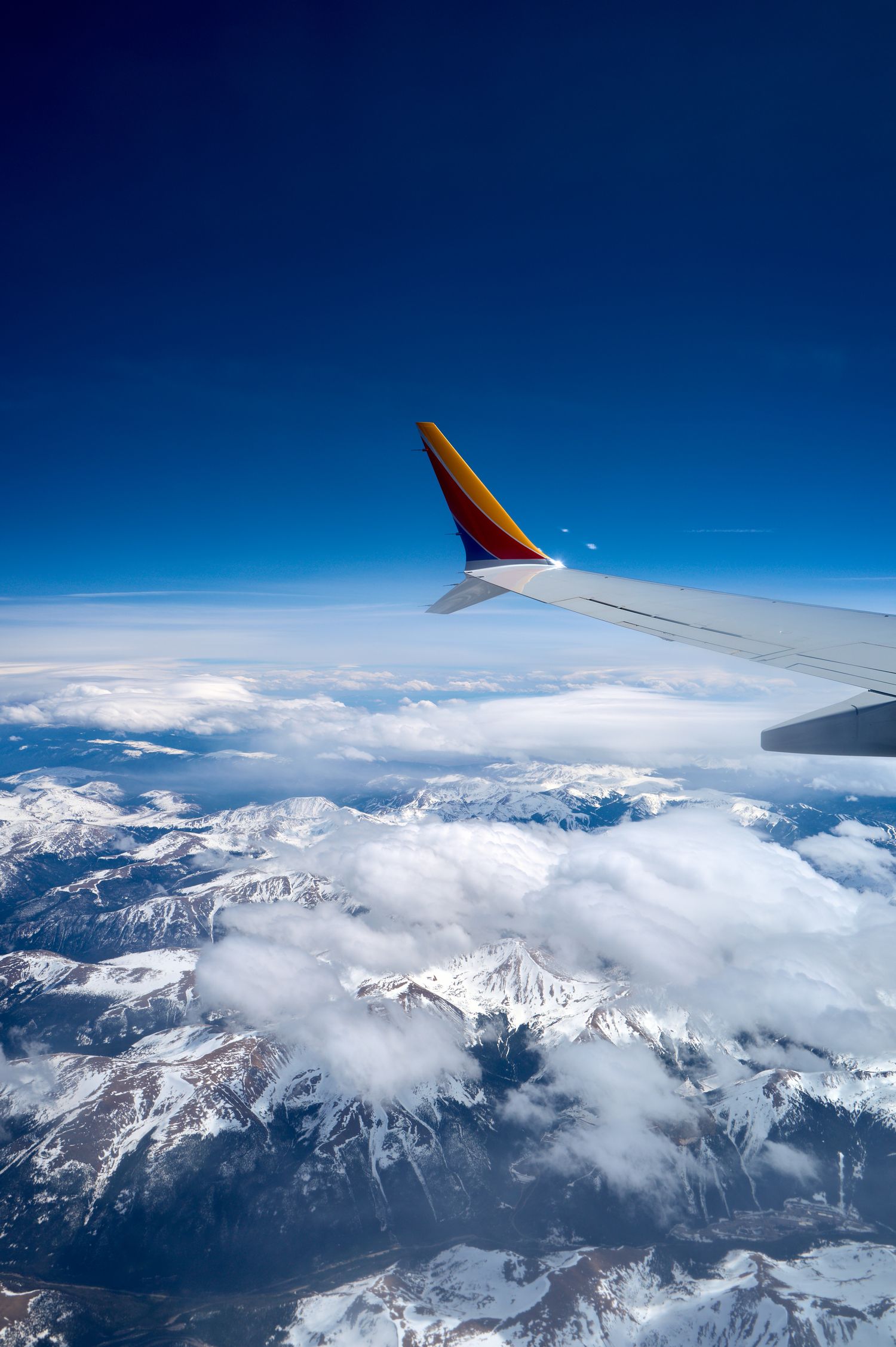 Airplane wing soaring over snow-capped mountains and clouds against deep blue sky.
