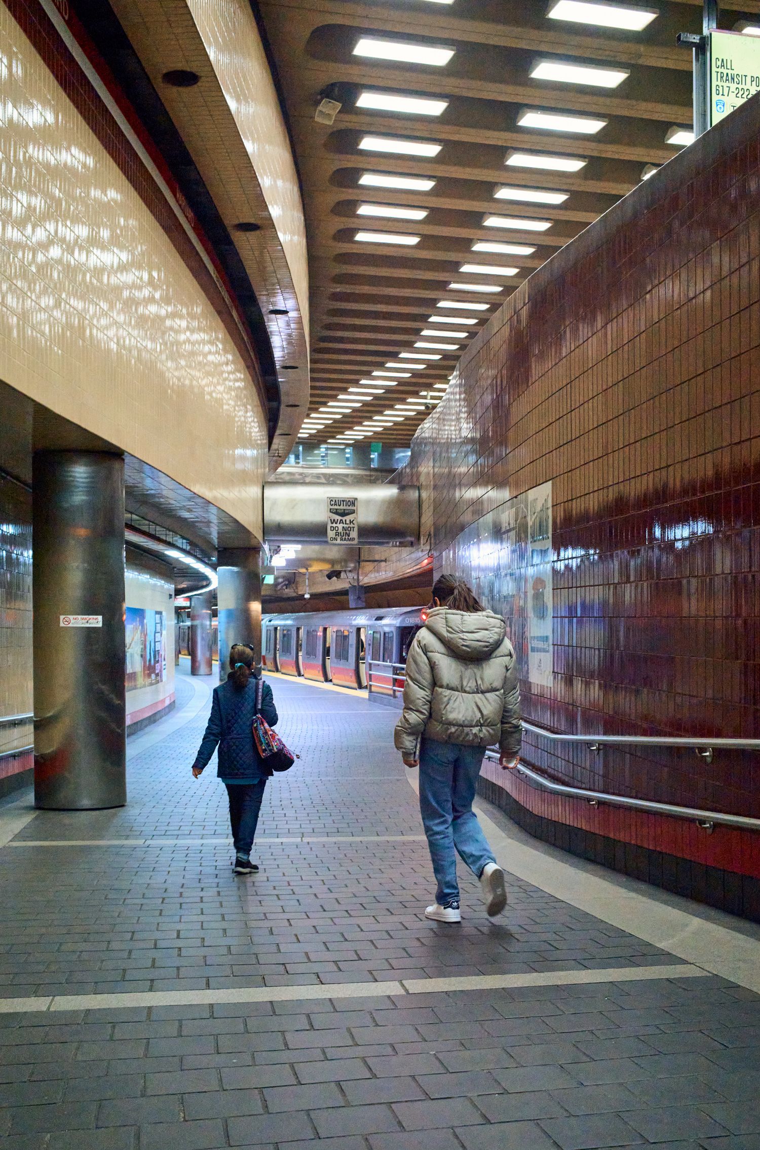 Modern subway station corridor with curved walls, fluorescent lighting, and commuters walking through.