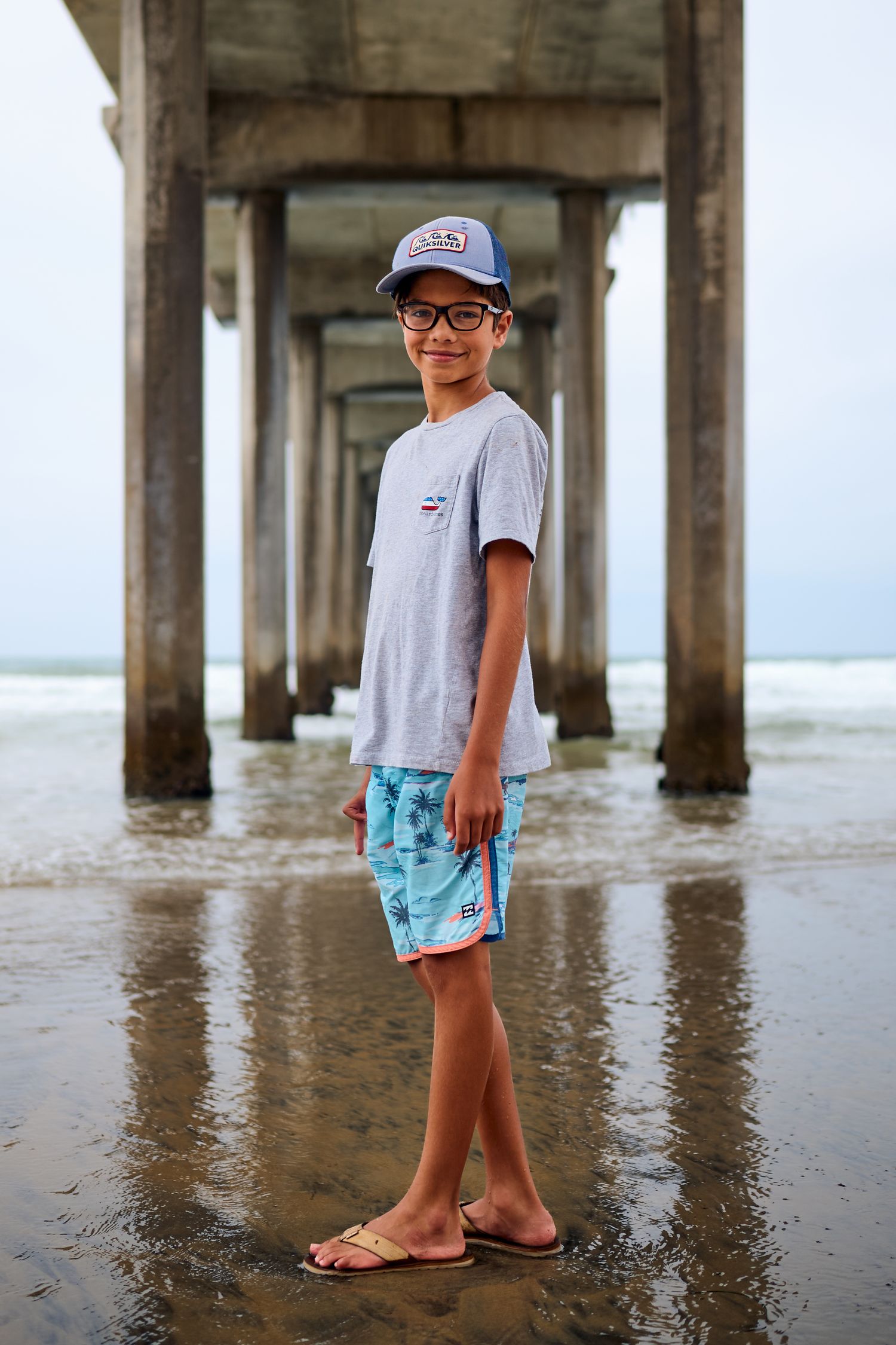 A young person wearing a baseball cap and swim trunks stands beneath a concrete pier at the beach.