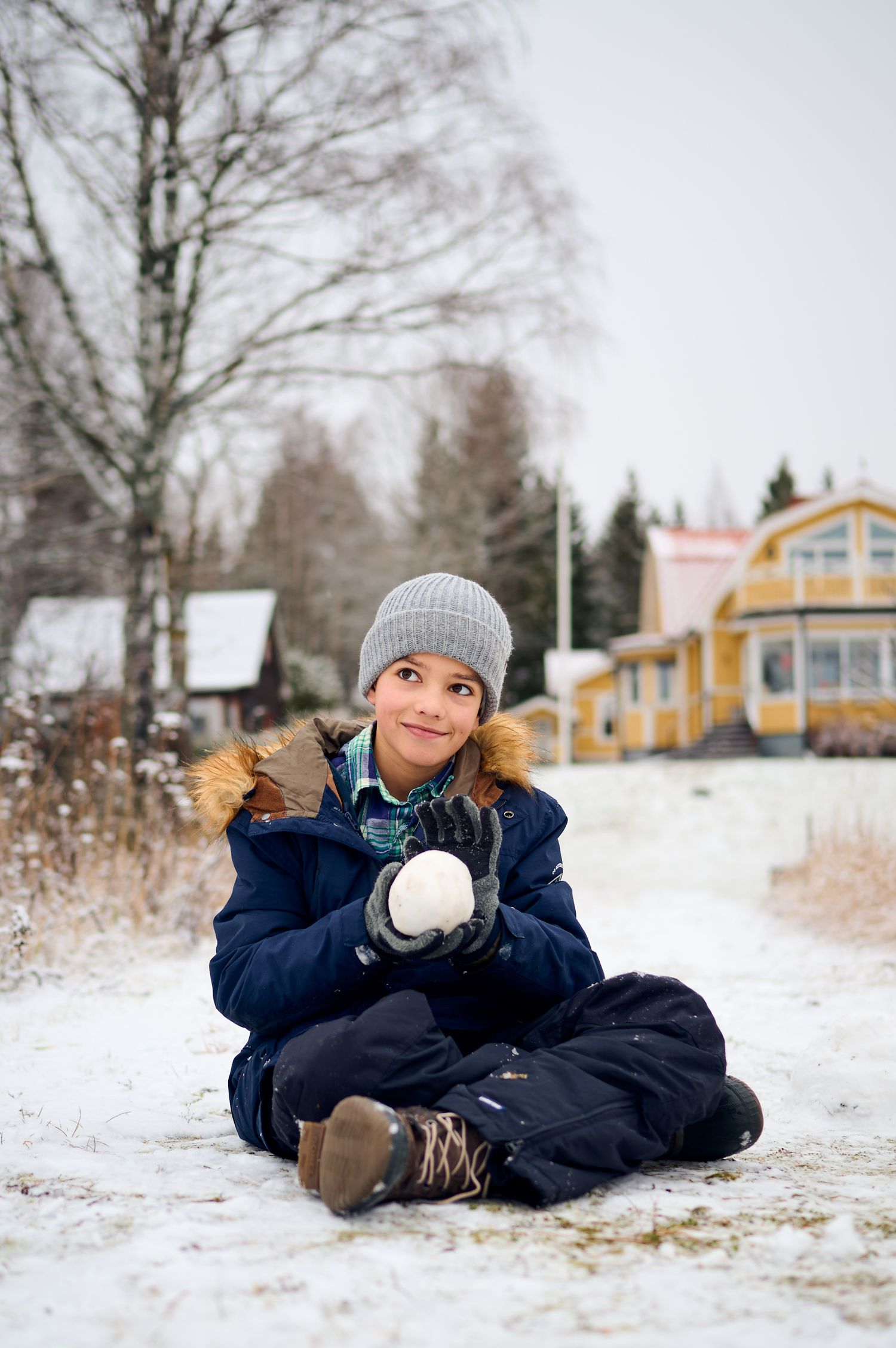 Person in winter coat and beanie sitting in snow holding a snowball.