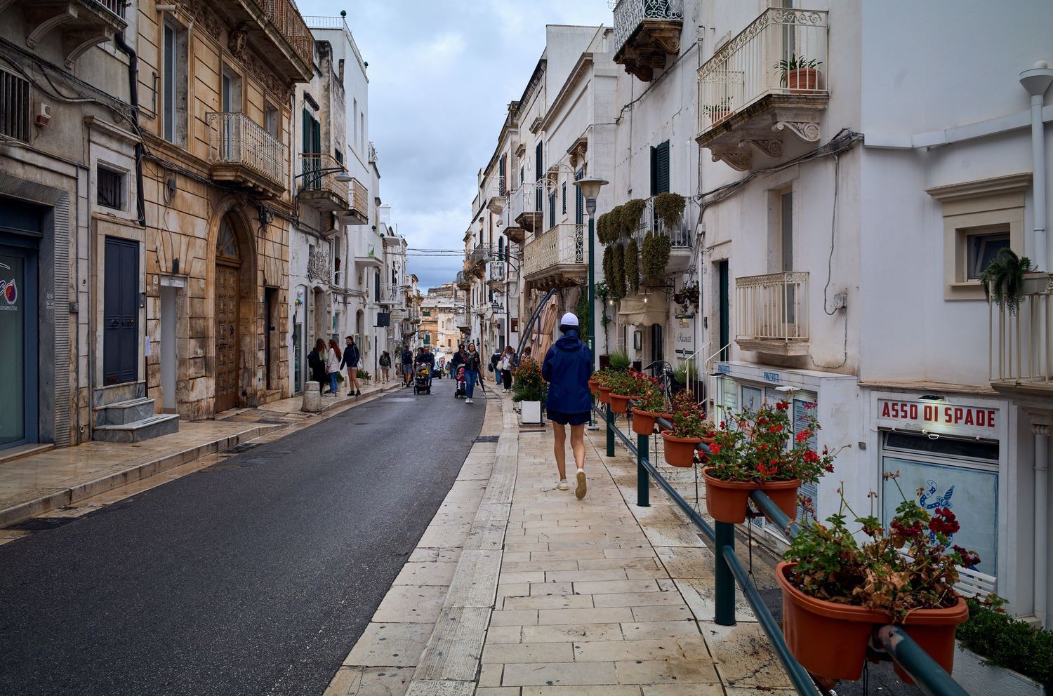 People stroll down a charming cobblestone street lined with historic buildings and potted flowers in a Mediterranean town.