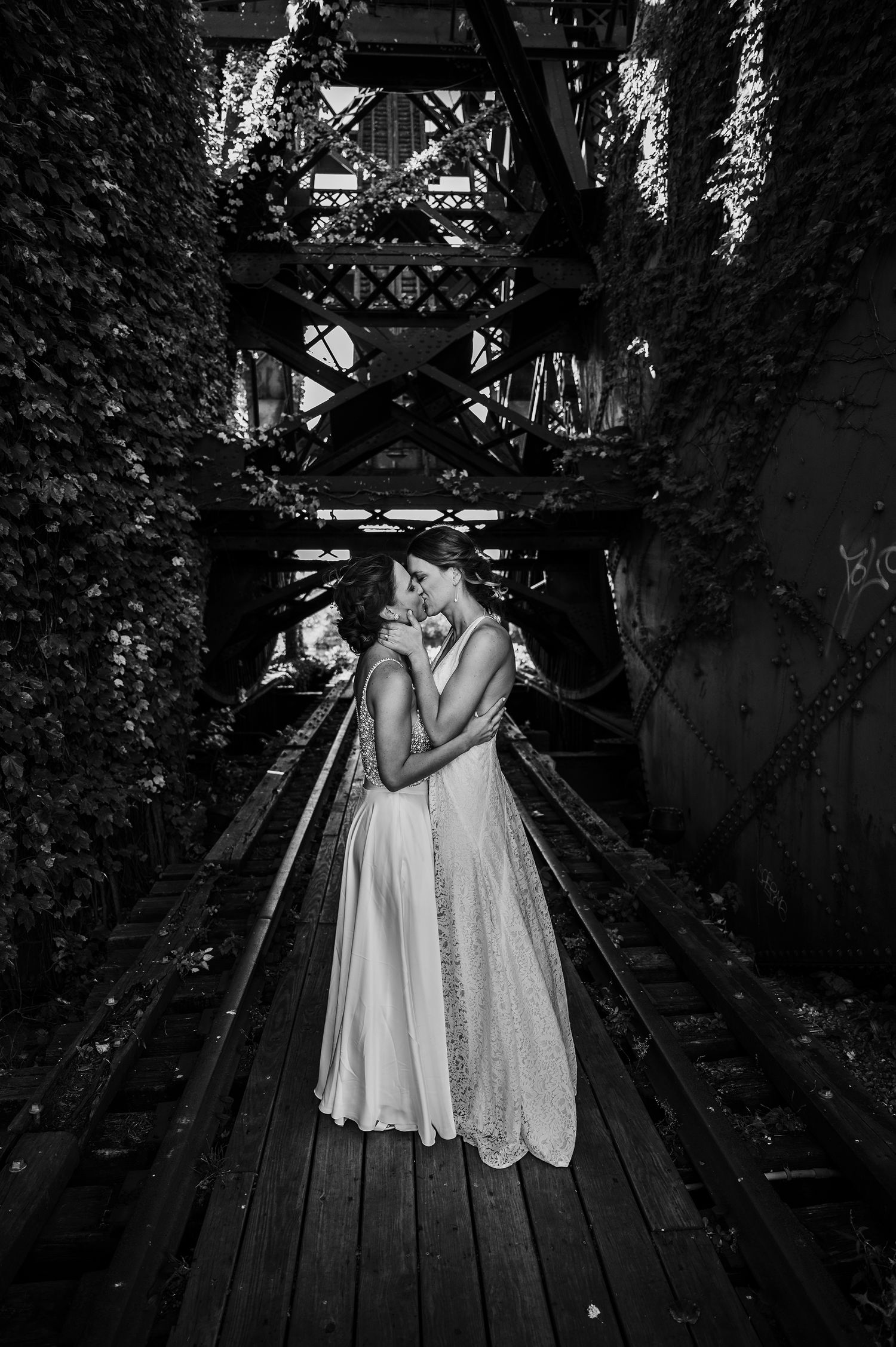 Black and white photo of a couple embracing on a wooden bridge surrounded by lush greenery and architectural beams.