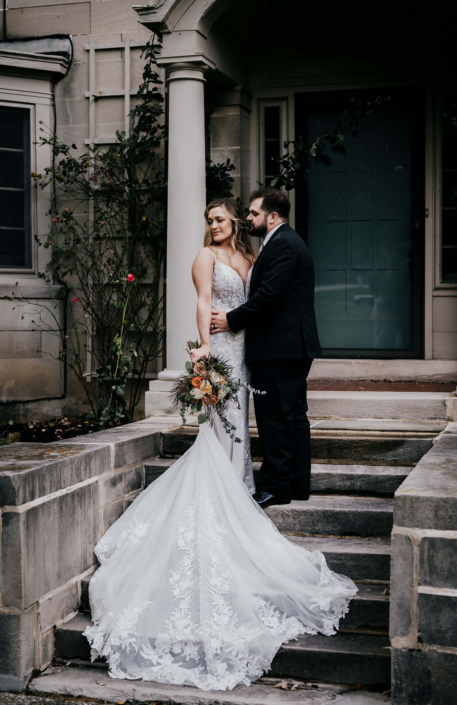 A wedding couple stands on stone steps outside a historic building with white columns and a trailing lace bridal gown.