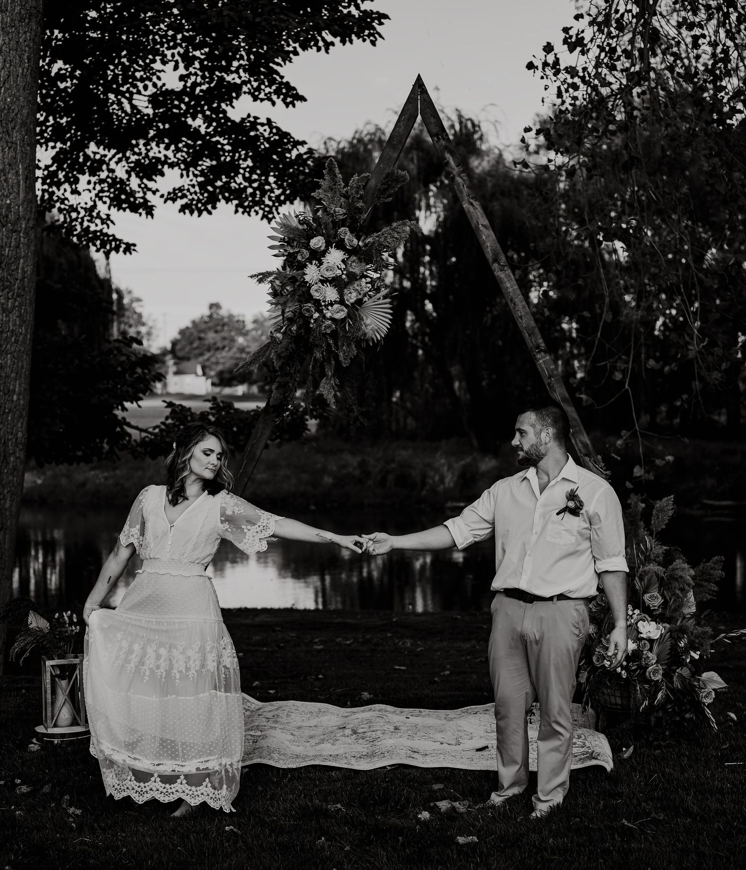 Two people hold hands in front of a triangular outdoor structure at a garden wedding ceremony.