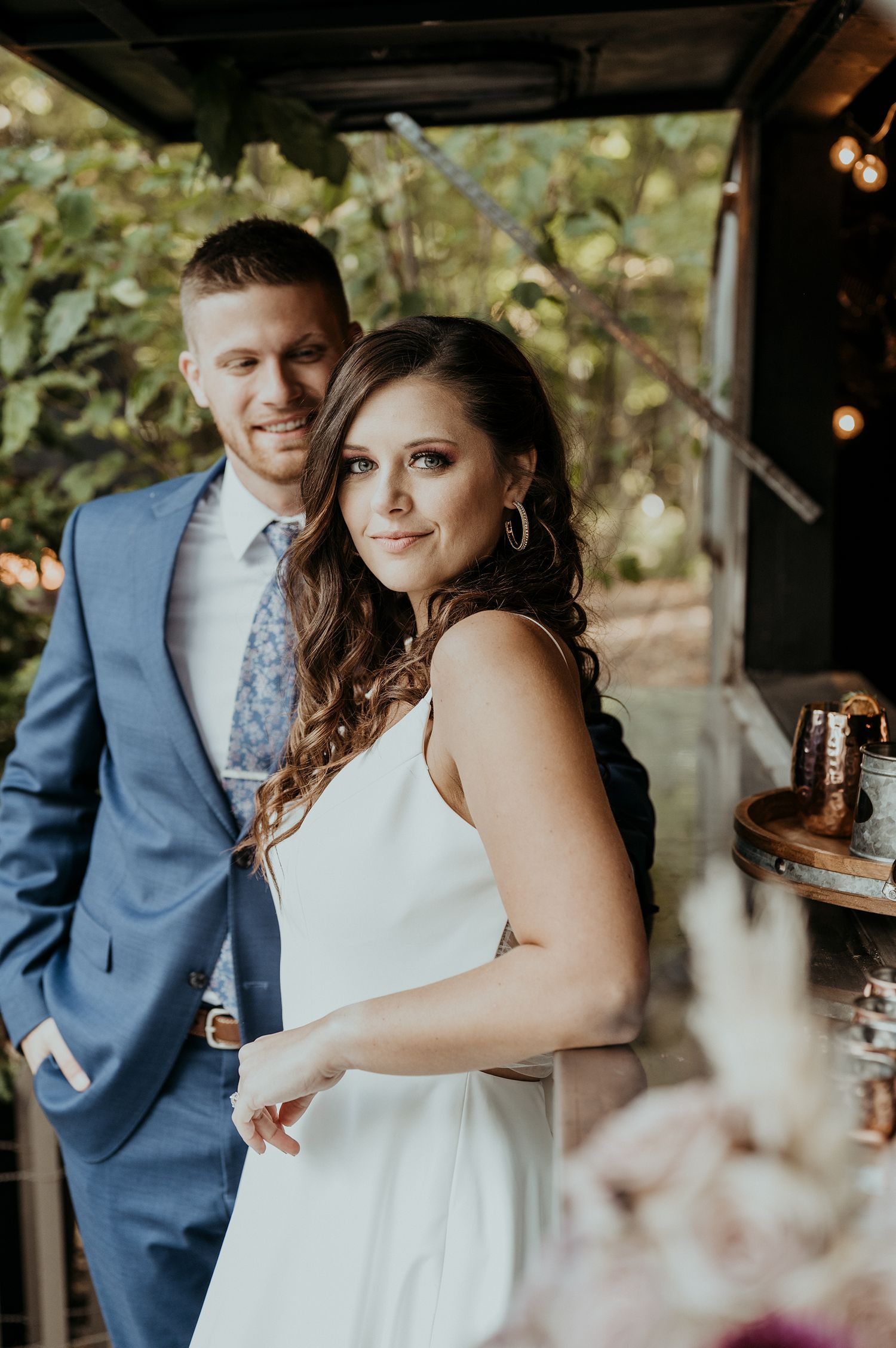 A couple in wedding attire pose under a covered outdoor area with string lights and natural greenery in the background.
