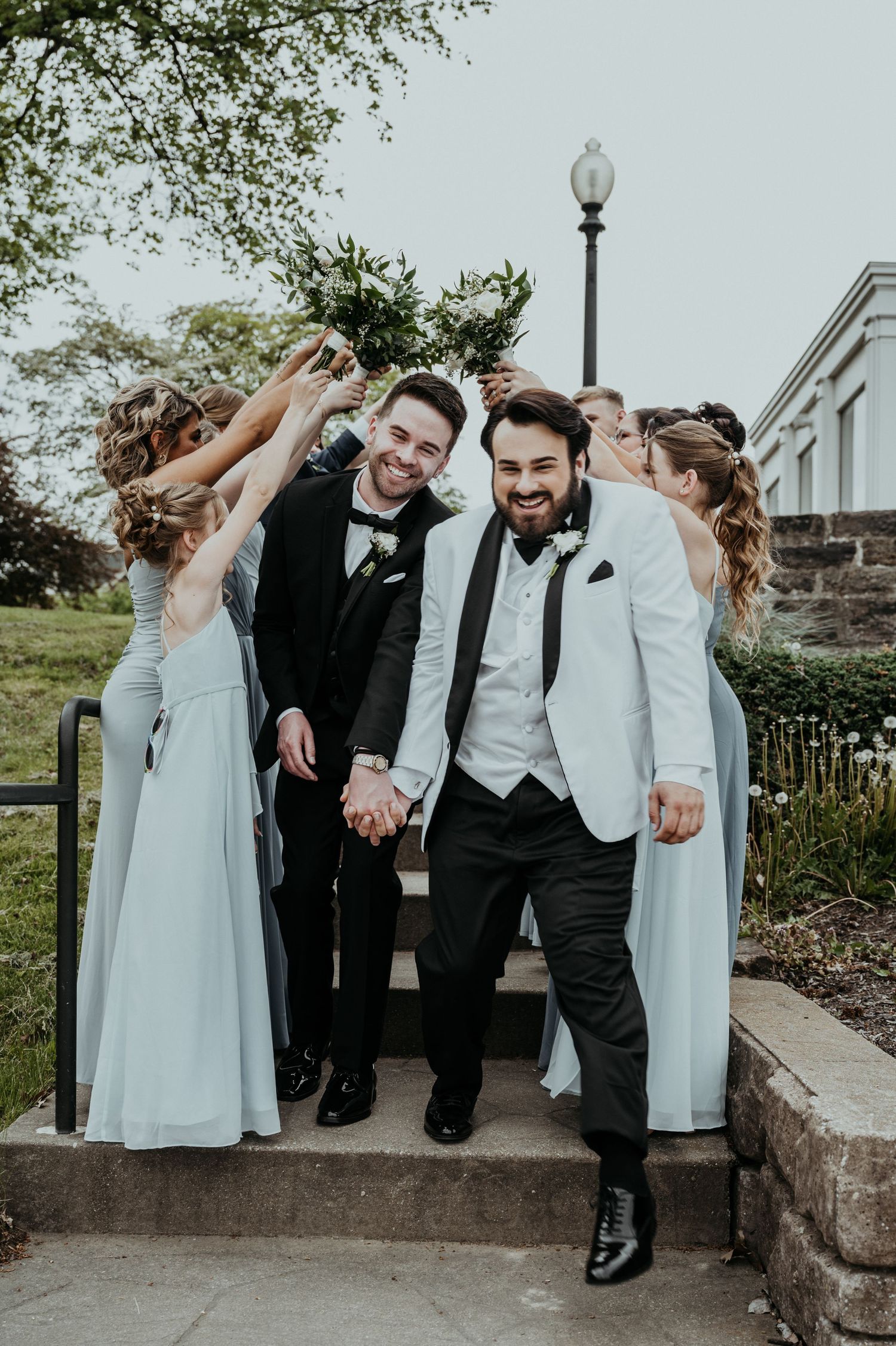 A wedding party dressed in black and light blue poses playfully on stone steps outdoors.