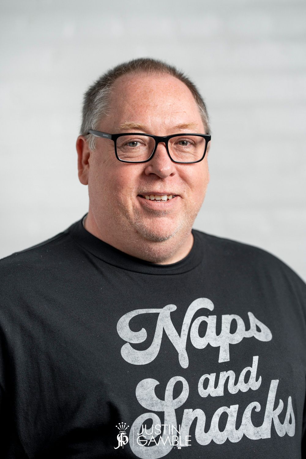 Portrait against light background wearing black t-shirt with 'Naps and Snacks' text and black-framed glasses.