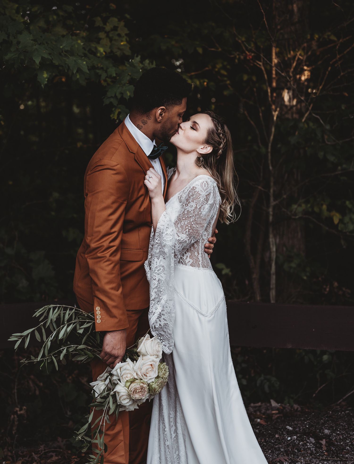 A romantic wedding couple in formal attire share intimate moments in a wooded outdoor setting at dusk.