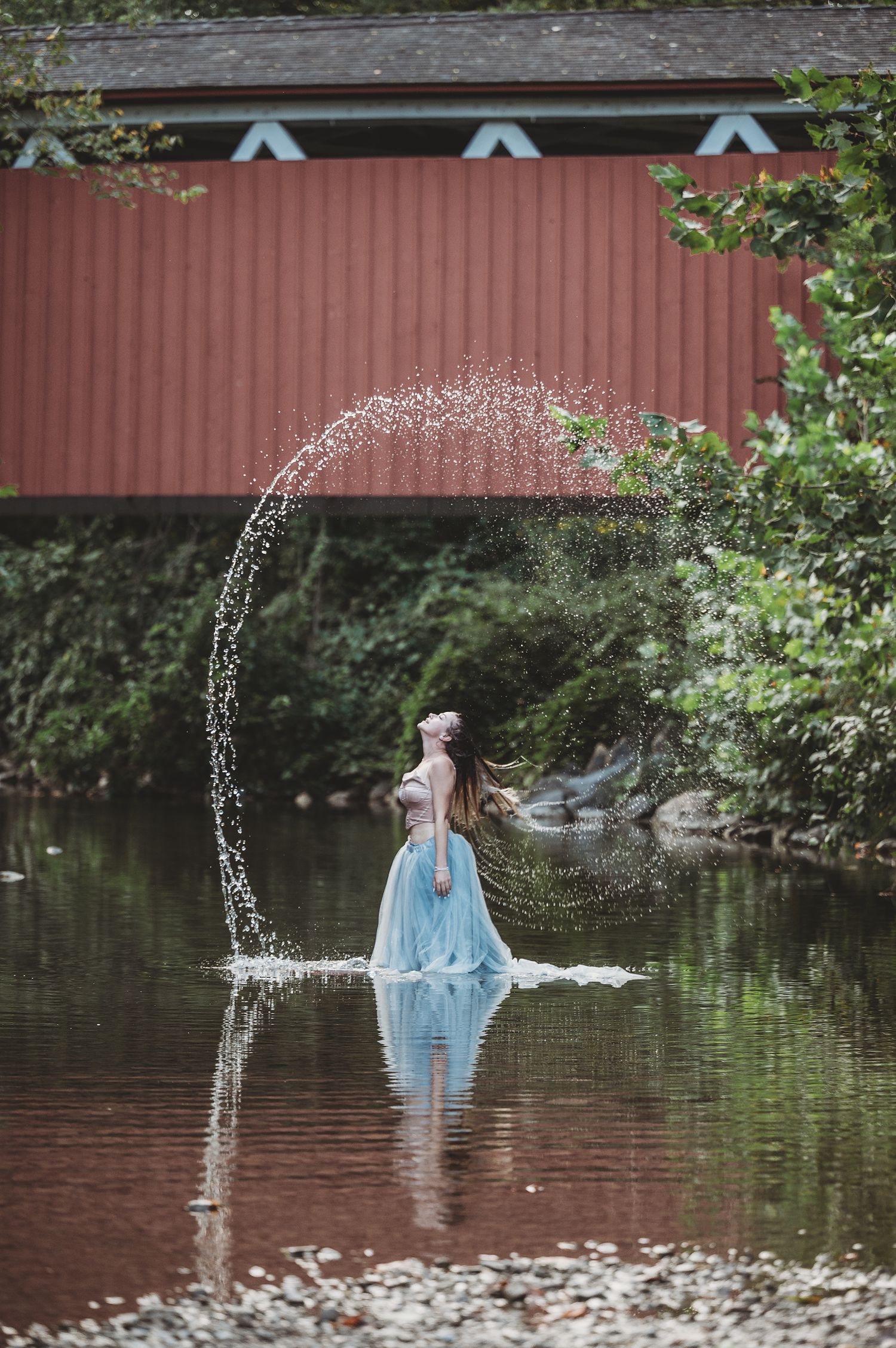 A figure in a flowing blue dress stands in shallow water under a red covered bridge as an arc of water sprays overhead.