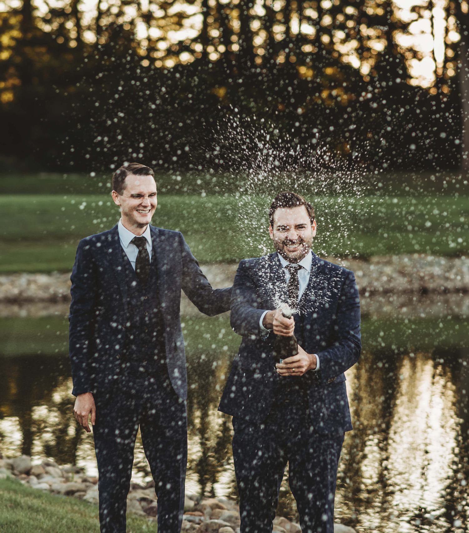 Two men wearing suits spray champagne outdoors at sunset near a reflective pond during a celebratory moment.
