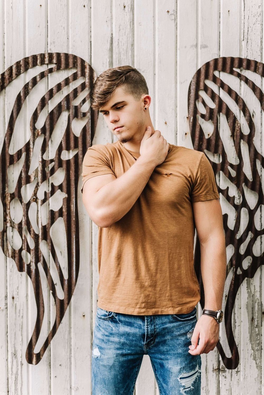 Someone in a brown t-shirt and ripped jeans poses against a white wall with painted black angel wings mural artwork.
