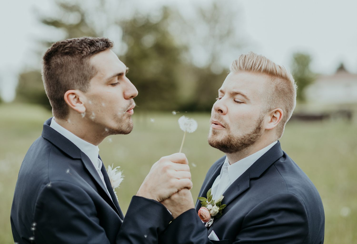 Two people in black suits share a dandelion in a romantic outdoor moment during a wedding celebration.