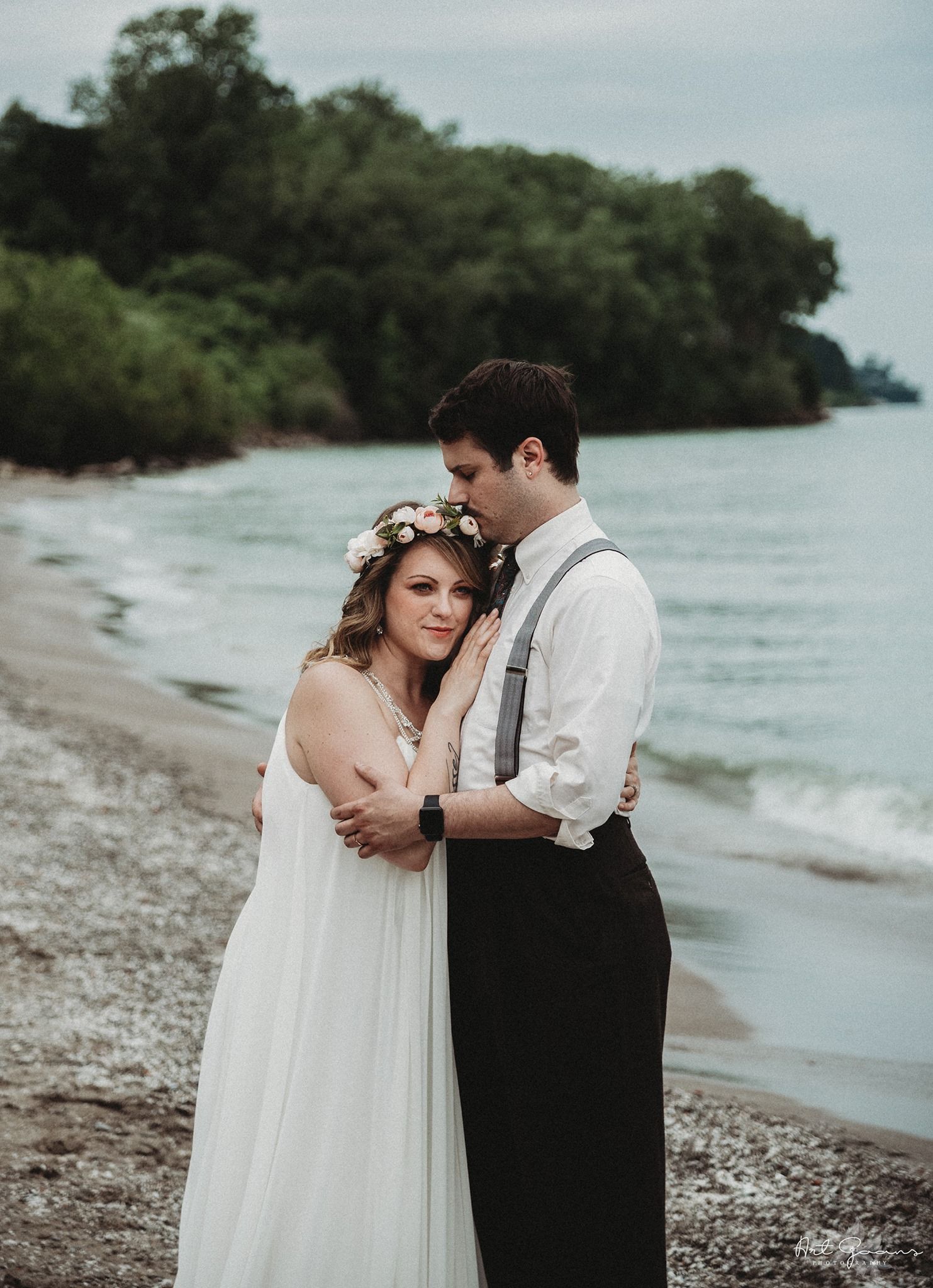 Couple shares romantic moment on a rocky beach shoreline with lush greenery and water in the background.