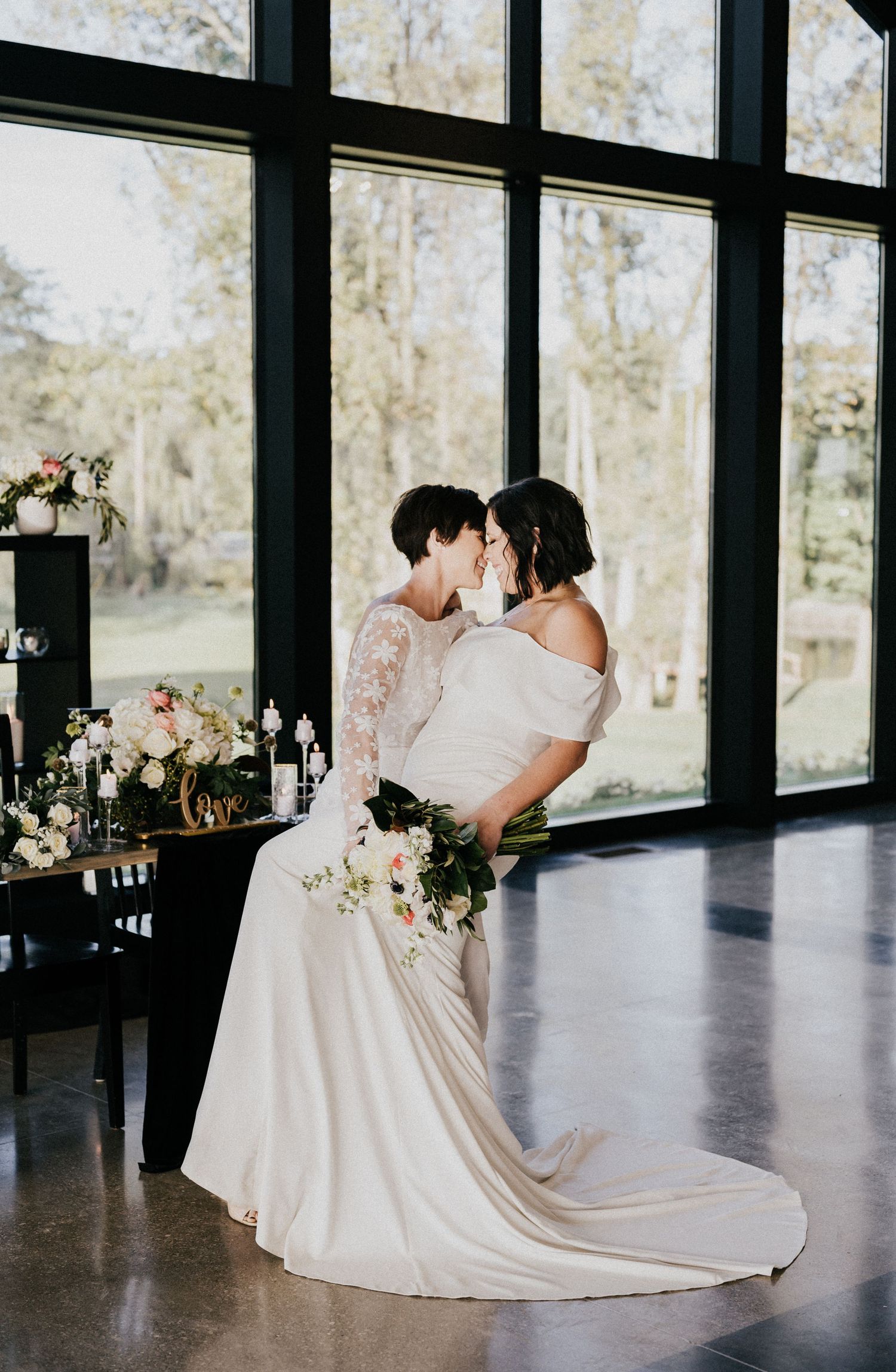 Two people share an intimate moment in elegant wedding attire by a large window overlooking nature.
