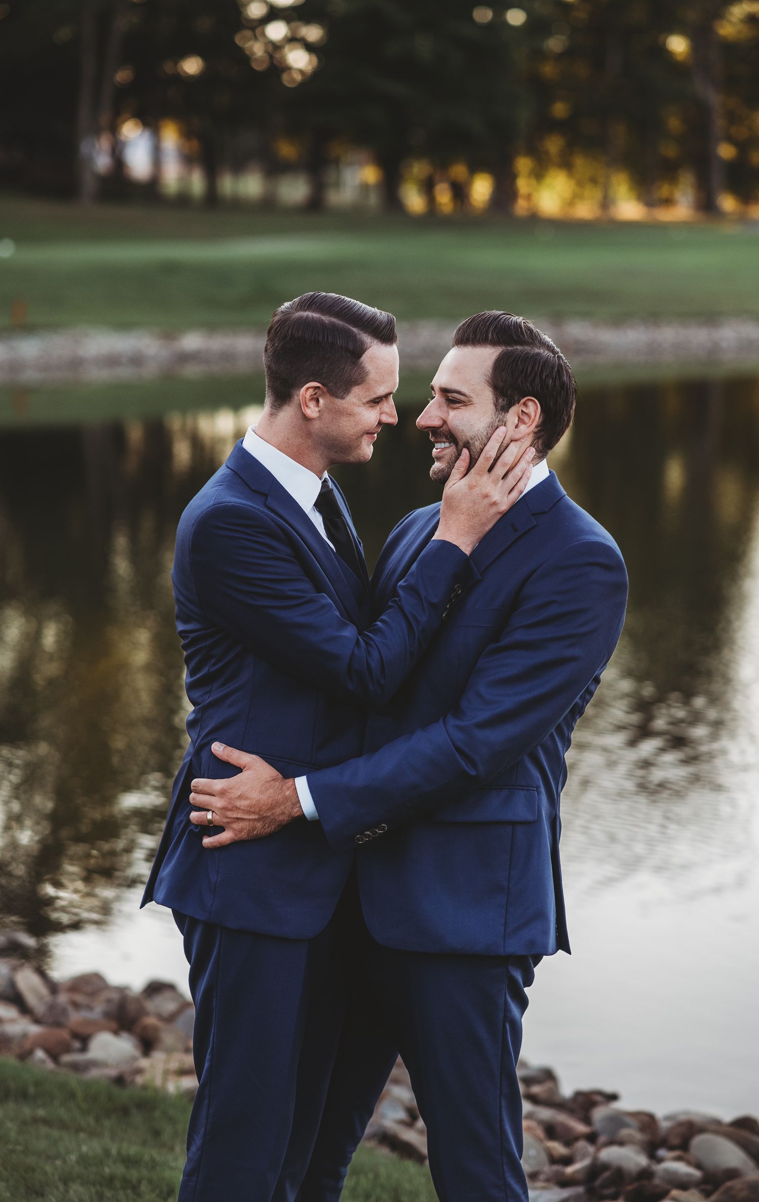 Two people in matching navy suits embrace near a tranquil lake at sunset.