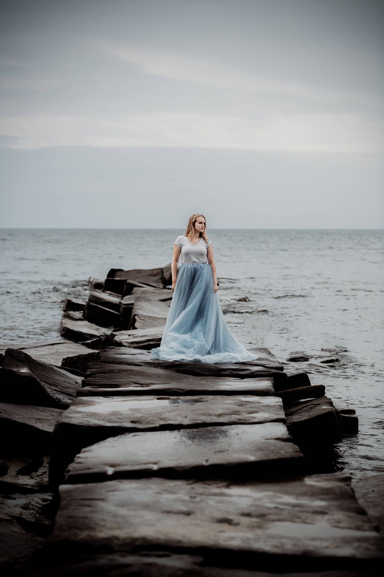 Ethereal figure in flowing blue dress poses along dramatic rocky coastline against moody gray ocean and sky.