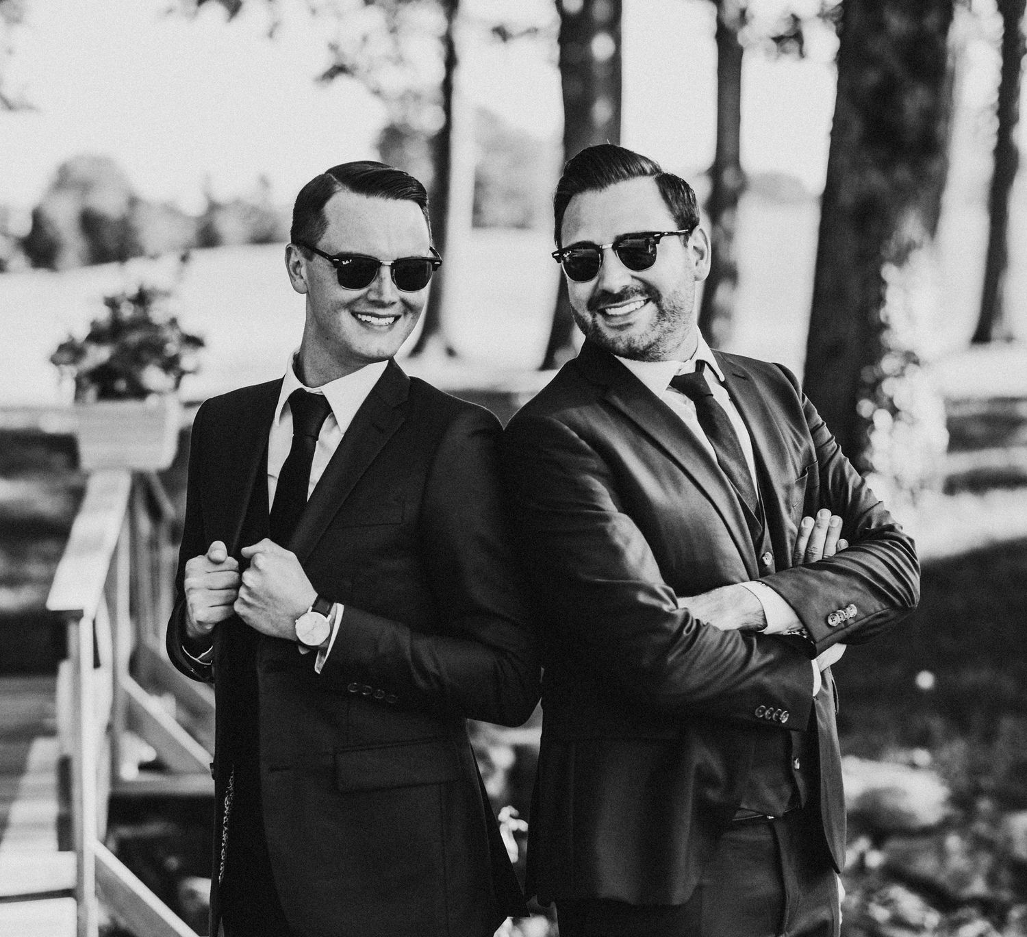 Two sharply-dressed men in black suits and sunglasses pose confidently outdoors in a black and white photo.