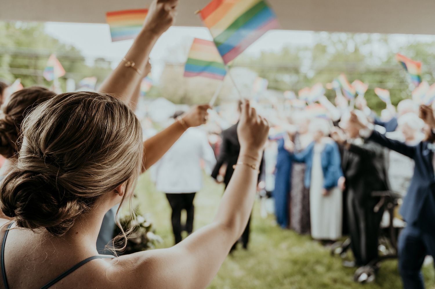 Wedding guests wave rainbow pride flags while celebrating with the newlyweds in an outdoor ceremony.