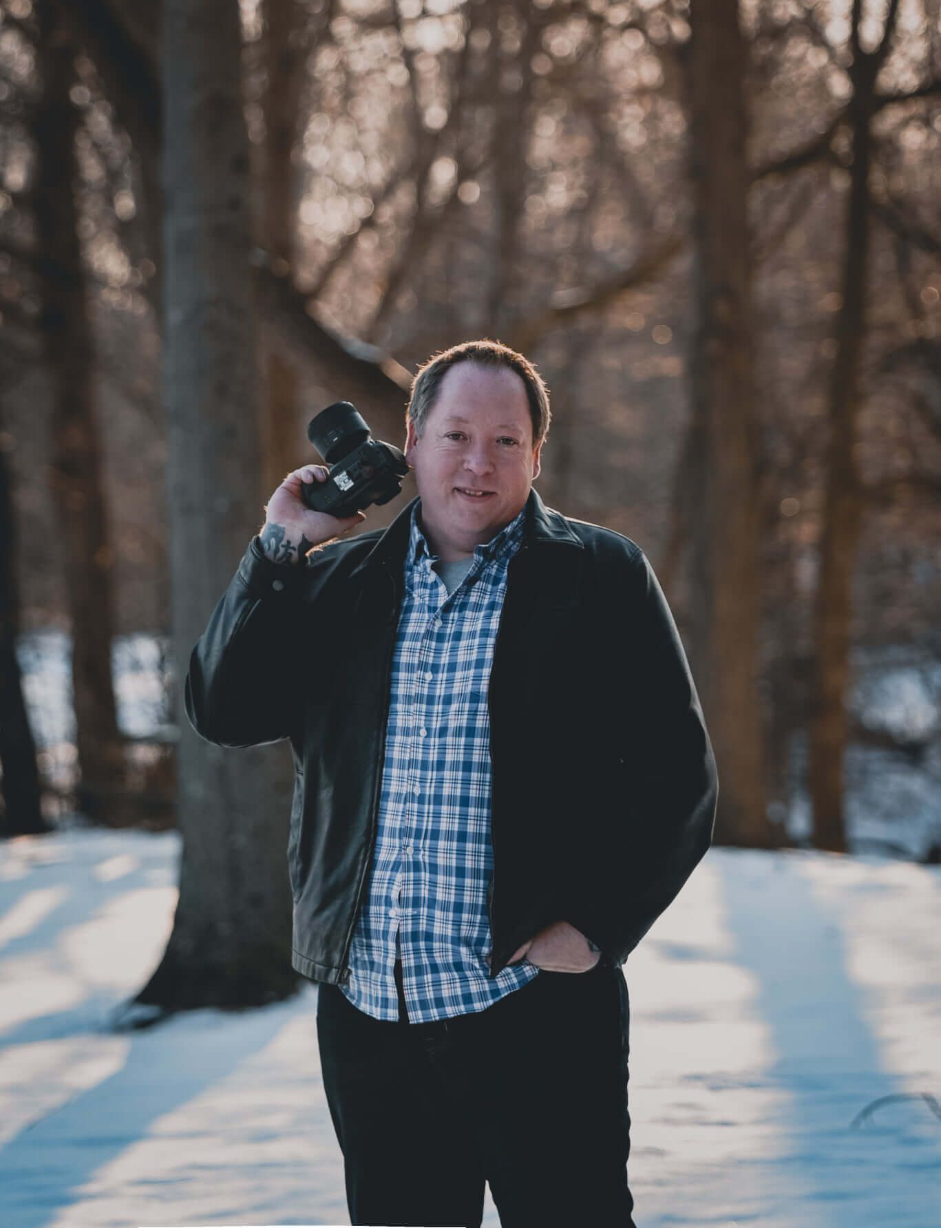 Person in plaid shirt and dark jacket walking through snowy wooded setting while throwing snow in winter landscape.