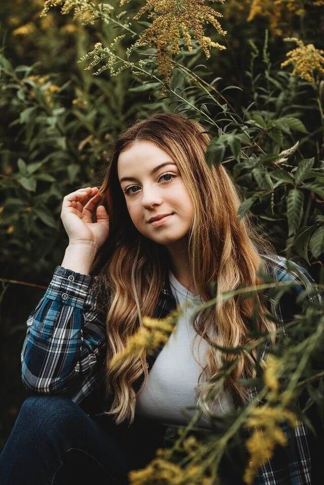 Portrait shot with long wavy hair and plaid shirt sitting among yellow flowering plants in natural outdoor setting.